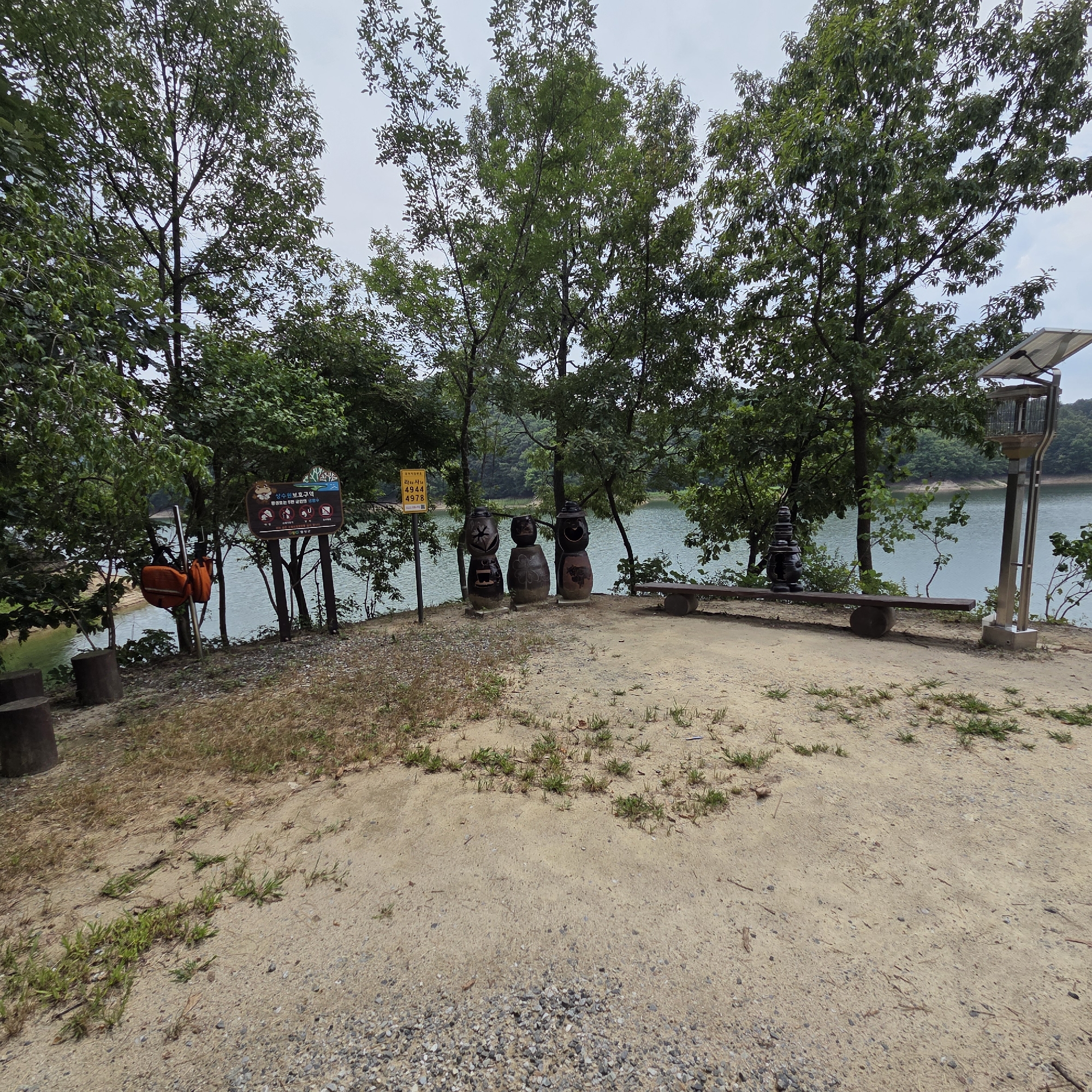 This is a photograph taken at the Gapcheon-myeon reservoir in South Korea. The foreground is a dirt path with scattered gravel and patches of grass. To the left, there are three dark, cylindrical tree stumps. In the middle ground, a body of water, likely the reservoir, is visible, bordered by trees. Several decorative ceramic figures resembling jars, some with faces, stand on the left side of the path. Behind them, an orange life preserver hangs on a pole. A wooden bench supported by stone blocks is situated in front of the reservoir, to the right of the ceramic figures. Further to the right, a solar-powered lamp post stands. Several trees with green foliage line the background, with a slightly overcast sky above. Two signs are posted on poles near the ceramic figures and the reservoir's edge. One sign has Korean text and images, and the other is a yellow sign with Korean text and numbers.