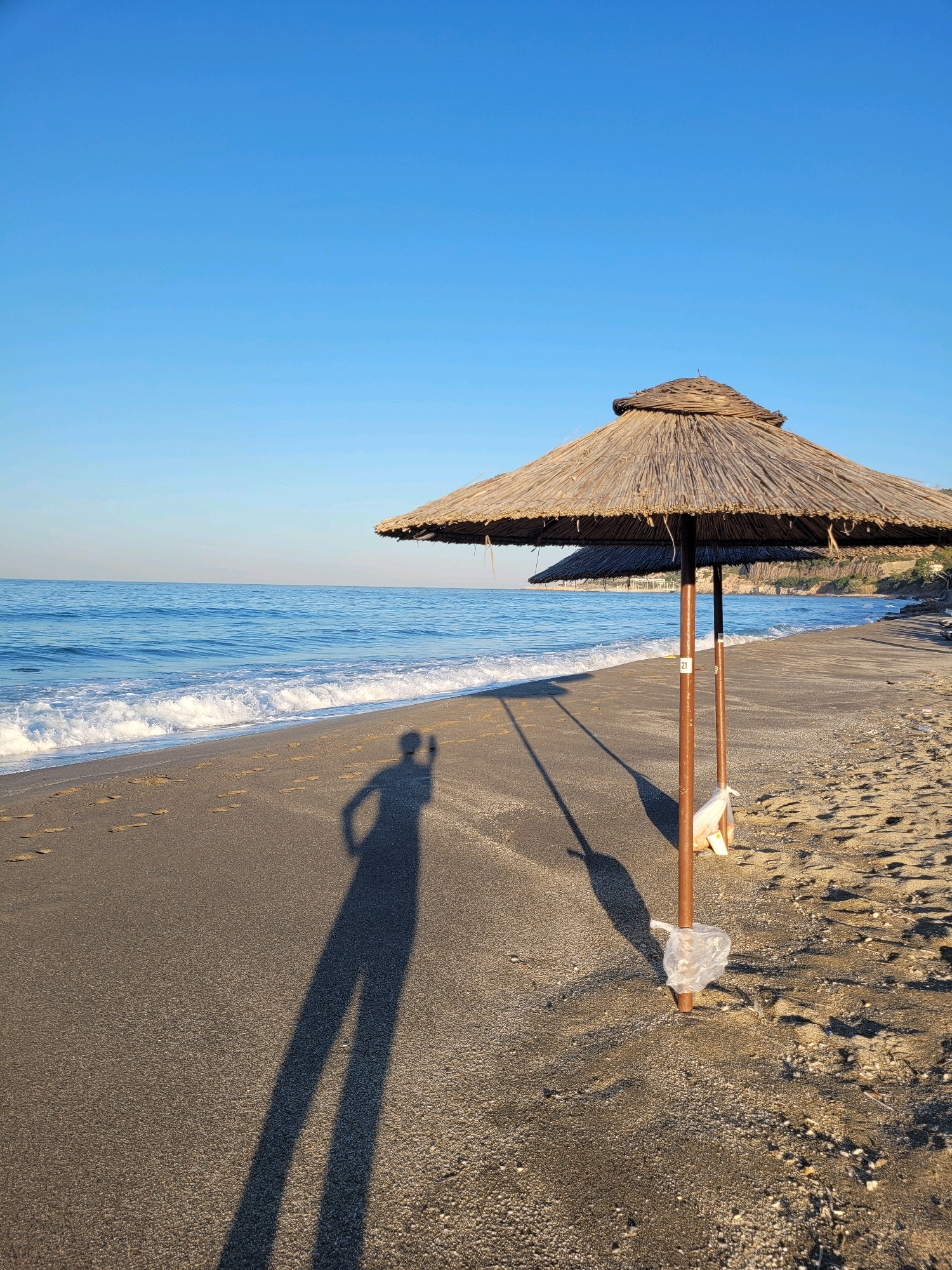 The image depicts a serene beach scene under a clear blue sky. In the foreground, a thatched umbrella stands on a sandy beach, casting a long shadow. To the left, gentle waves of the Mediterranean Sea lap onto the shore, leaving ripples and wet sand. The beach is mostly empty, with only the shadow of a person holding a phone visible, suggesting they are taking the photograph. The overall impression is one of tranquility and a quiet morning or late afternoon on the coast. It is difficult to pinpoint the exact location within Alanya, Turkey, without further information, but it is consistent with the numerous beaches along its coastline.