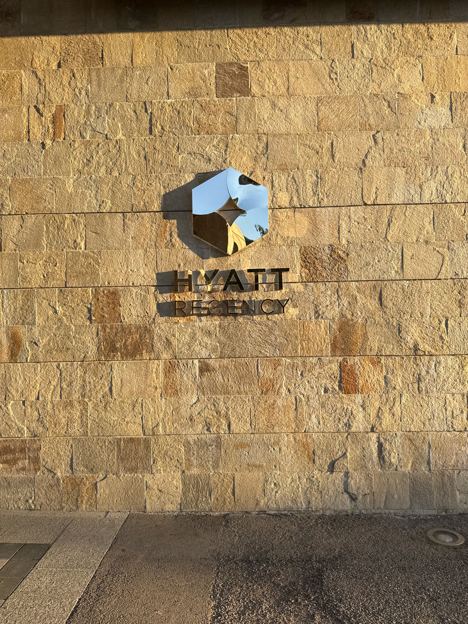 A close-up, eye-level, vertical shot showcases the exterior wall of the Hyatt Regency hotel in Razliv, Bulgaria. The wall is constructed of large, rough-hewn, tan-colored stone blocks, with visible mortar lines. Sunlight casts long shadows across the textured surface, highlighting the stone's imperfections and creating a warm, golden hue. Centered on the wall is the Hyatt Regency logo, a stylized silver hexagon with a smaller, lighter hexagon inside. Below the logo, the words "HYATT REGENCY" are spelled out in dark, metallic letters. The bottom of the frame shows a sidewalk made of gray stone tiles and a dark, gravelly area, possibly a pathway or landscaping. A circular, recessed light fixture is visible in the lower right corner of the frame. The scene appears to be a clear, sunny day, with the late afternoon sun bathing the facade.