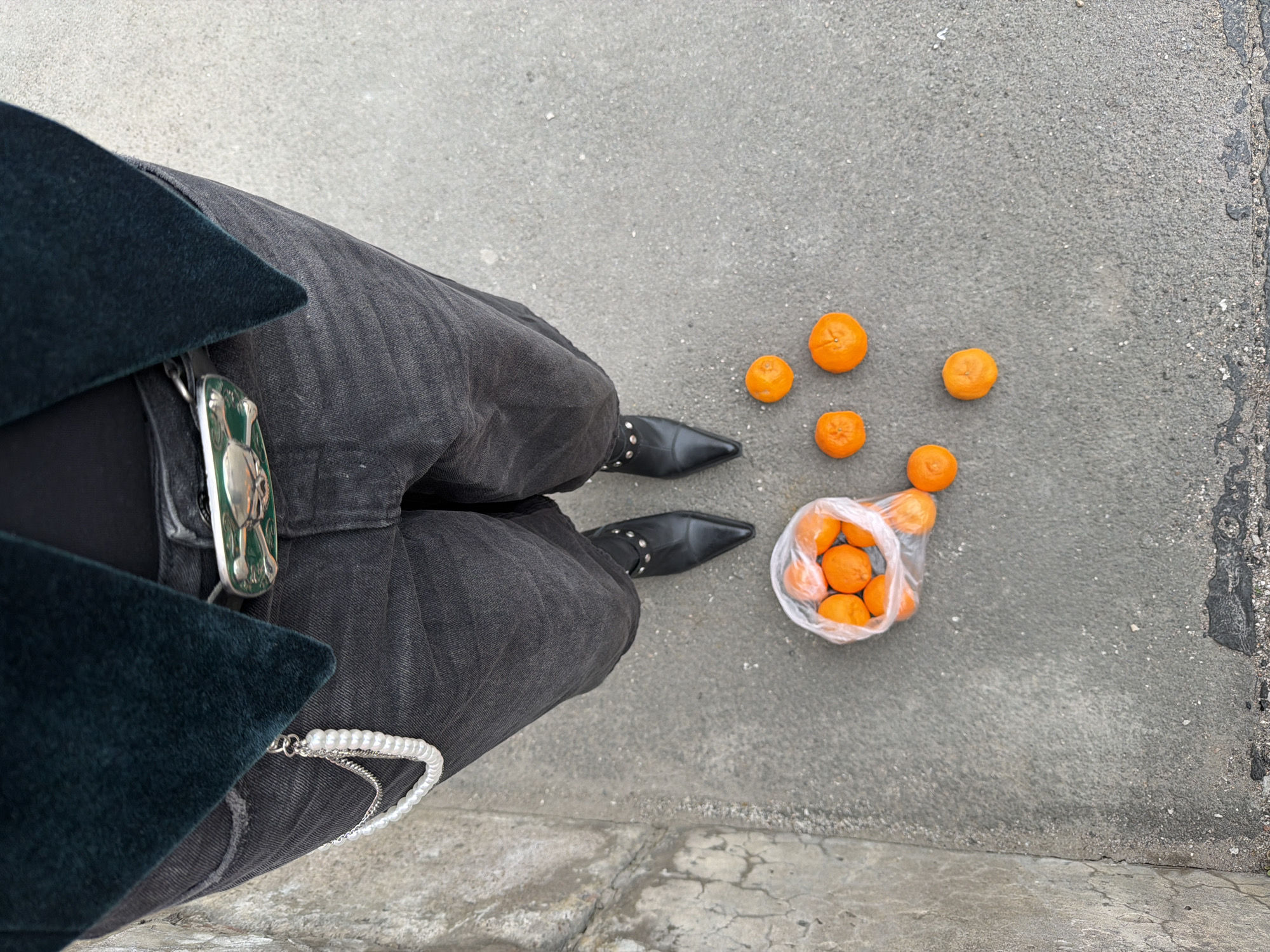 A person stands on a cracked grey concrete surface, possibly a sidewalk, in Saint Petersburg, Russia. They are wearing dark grey jeans, black leather boots with small silver studs around the ankles, a black top, and a dark green jacket. A silver belt buckle with a green design and a string of pearls adorn the jeans. To the right of the person, partially obscured by the hem of the jeans, a clear plastic bag contains several oranges. Scattered around the bag are six more oranges. The background is entirely the concrete surface.