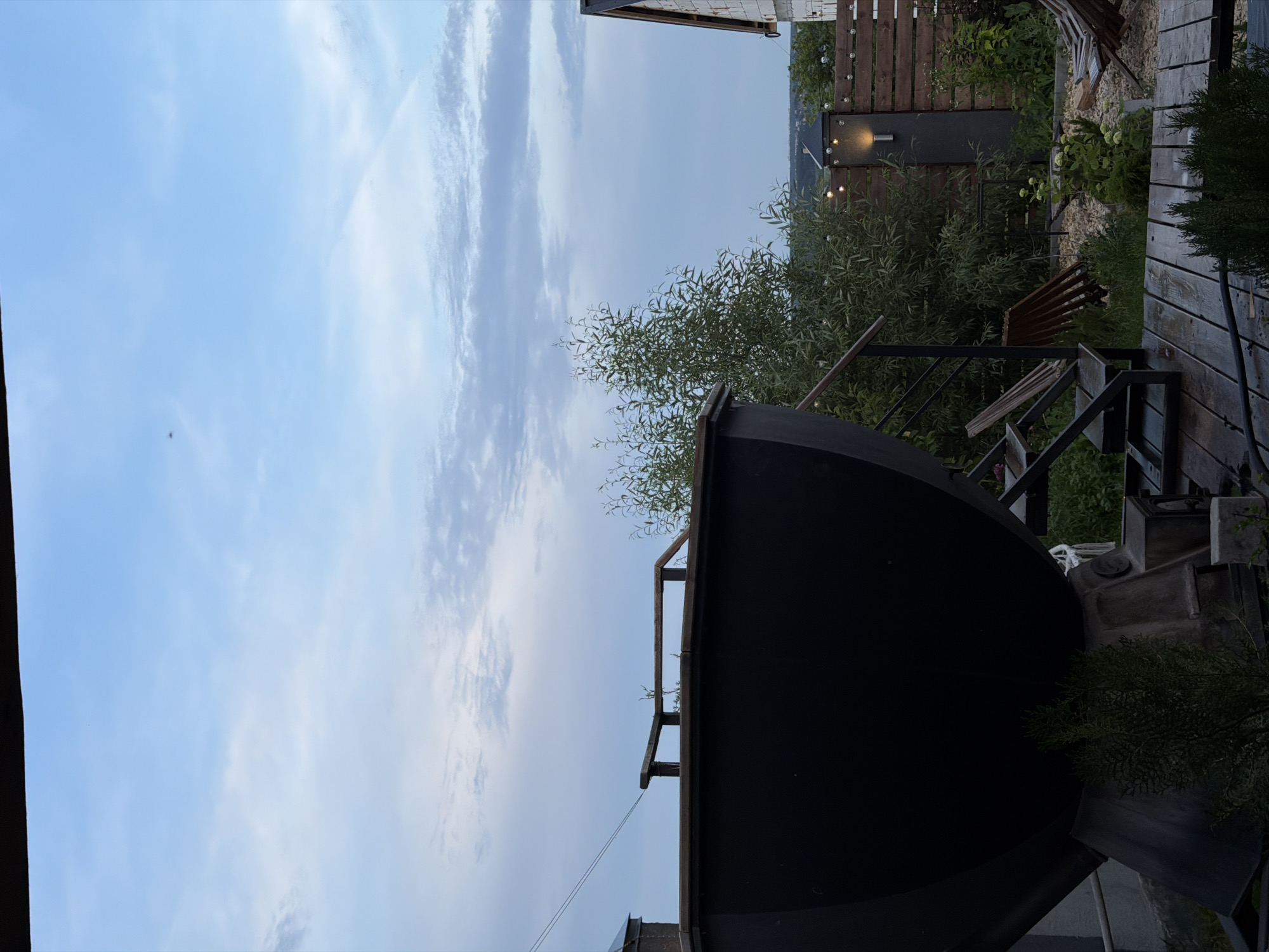 The image depicts an outdoor scene in a residential yard, possibly in the city of Semiluki, Russia. In the foreground, a large, dark, semi-spherical object, which appears to be a large cauldron or a decorative water feature, dominates the left side of the frame. Stairs made of metal and wood lead up to this object. The ground is covered with wooden planks and gravel, with various plants and greenery scattered throughout.
Behind the cauldron, a wooden fence with string lights is visible, separating the yard from what appears to be a neighboring property. A partially constructed building with exposed brickwork is visible on the right side of the fence. The background reveals a gently rolling landscape with trees and some distant structures under a pale blue sky with scattered clouds. The lighting suggests it is either early morning or late evening. There are no people visible in the image.