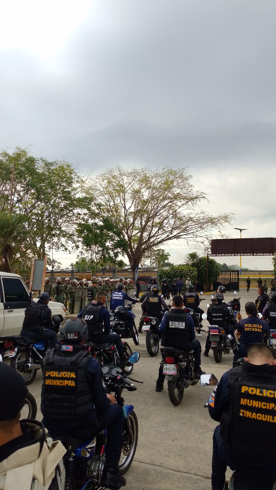 The image depicts a daytime gathering of multiple Venezuelan police forces and their vehicles in Tinaquillo, under an overcast sky. In the foreground, several police officers are seated on motorcycles, many facing away from the camera. They wear dark tactical vests, helmets, and dark uniforms.
Visible text on their vests identifies various units: "POLICIA MUNICIPAL," including specific markings for "POLICIA MUNICIPAL TINAQUILLO," as well as "POLICIA ESTADAL" (State Police). Other visible abbreviations include "DIP," "S.I.P," and "DCDO." One officer's helmet bears the text "5.11 TACTICAL GEAR." License plates such as "AM4D09K" are visible on the motorcycles.
Behind the motorcyclists, a larger group of officers or personnel is formed up on foot, appearing to be a riot control unit. They are equipped with helmets and carry large riot shields. This group is positioned near a white SUV and a fenced area or gate.
The background features numerous green trees, some with dense foliage, and distant structures, suggesting an outdoor, potentially semi-urban or administrative location. The scene conveys a sense of readiness, deployment, or preparation for a public order event involving coordinated efforts from different police agencies in Tinaquillo.