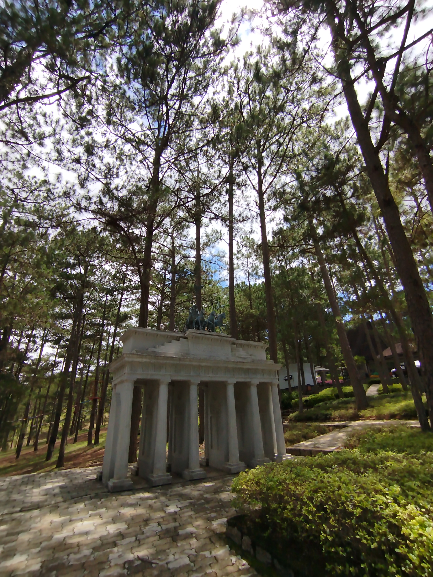 The image shows a monument in the forest area of Lam Vien, Da Lat, Vietnam. The monument is a rectangular structure with six columns and a statue of horses on top. It is surrounded by pine trees and green bushes. The ground is covered with patterned bricks. The sky is visible through the trees and is partly cloudy.