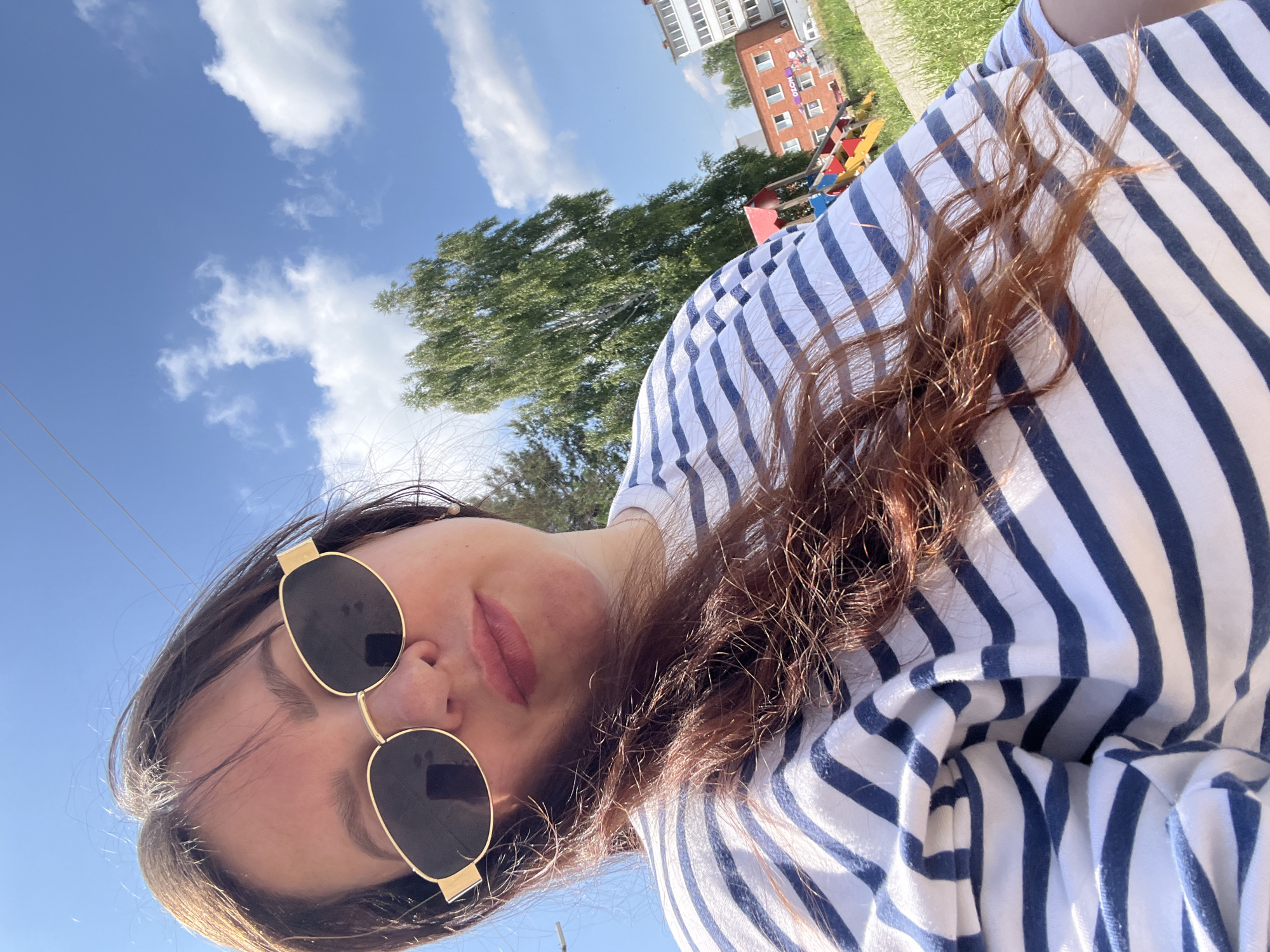 A selfie taken outdoors shows a woman wearing a blue and white striped t-shirt and gold-rimmed sunglasses. Her long, dark brown hair is partially covering her face. In the background, a green tree stands against a blue sky with scattered white clouds. To the right, a brick building and a playground structure are visible. The image appears to be taken in a residential area, likely in Noviy, Russia, judging by the architecture and typical landscape.