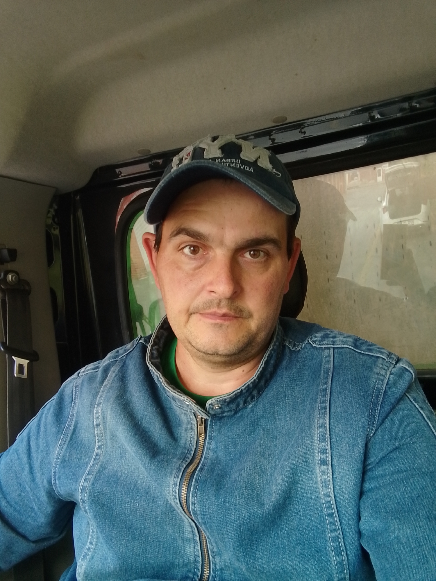 A man is seated inside what appears to be a vehicle, possibly a tractor or truck cab, looking directly at the camera. He is wearing a blue denim jacket over a green t-shirt and a blue baseball cap with white lettering. The interior of the vehicle is visible, including a seatbelt on the left side and a window in the background that reflects an outdoor scene, possibly a street with buildings and vehicles, suggesting an urban or suburban environment. The man's face is well-lit, and his expression is neutral. The overall lighting suggests it is daytime. There is no indication of the specific location in Wedel, Germany, within the image itself.