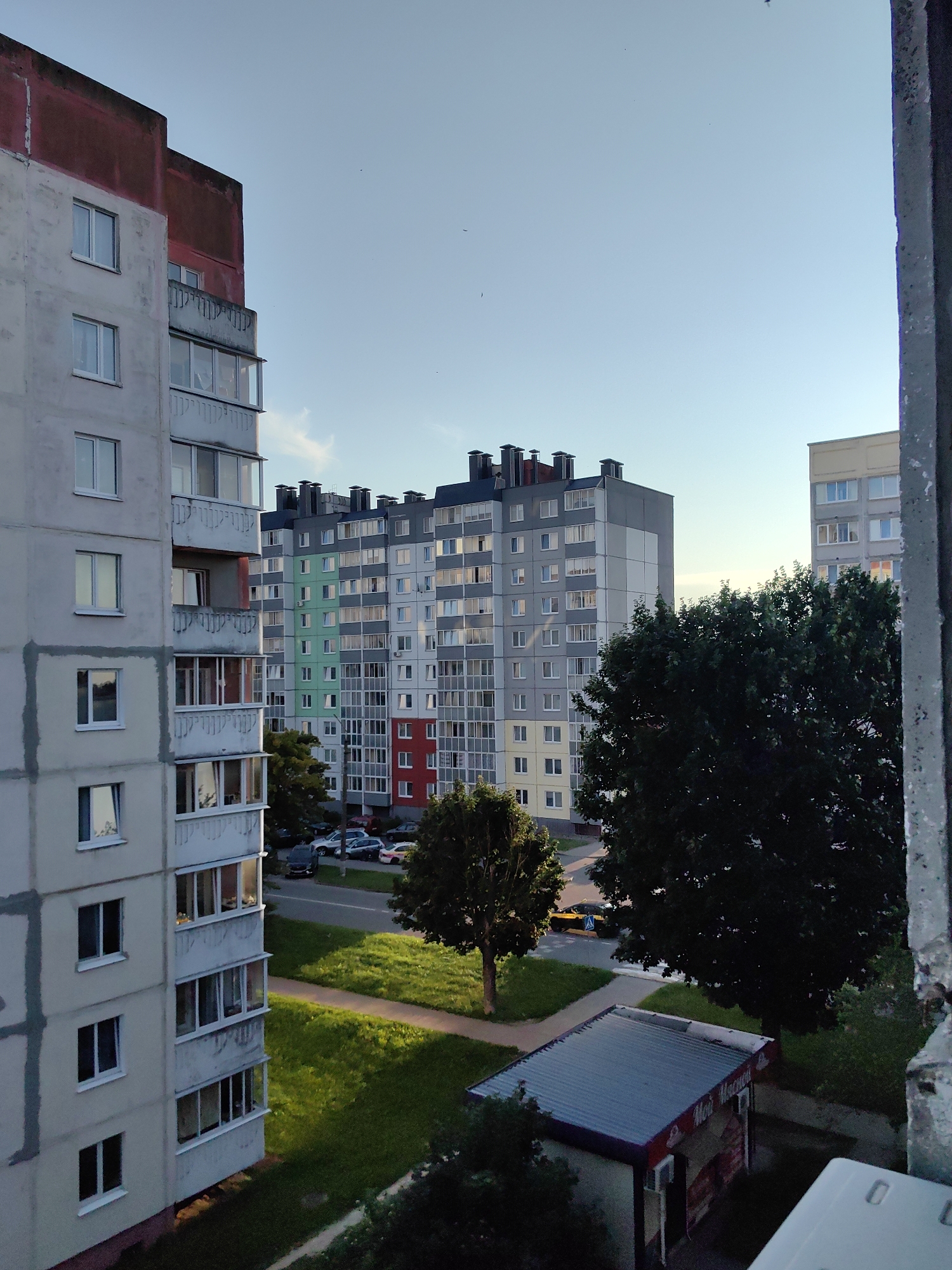 The image captures a view from a window overlooking a residential area in Fanipal, Belarus. In the foreground, a portion of a concrete window frame is visible on the right. To the left, a tall, light-colored apartment building with red accents at the top dominates the left side of the frame. It features multiple floors with evenly spaced windows and balconies.
Further into the scene, across a road and green lawn, stands another larger apartment building. This building is characterized by its colorful facade, with sections of green, red, and yellow interspersed with grey and white. The building has numerous windows and dark ventilation units on its roof. To the right of this, another similar apartment building, primarily grey and white, is visible.
A road runs between the apartment buildings and the grassy area. Several cars are parked along the road. Two mature trees are situated in the middle ground, one to the left of the colorful apartment building and another, larger, darker tree to the right.
In the lower right corner, a small, single-story kiosk with a corrugated metal roof is visible. It appears to be a shop, with signage in Cyrillic script. The sky is clear and light blue, indicating either early morning or late afternoon. The overall impression is of a typical urban residential environment.