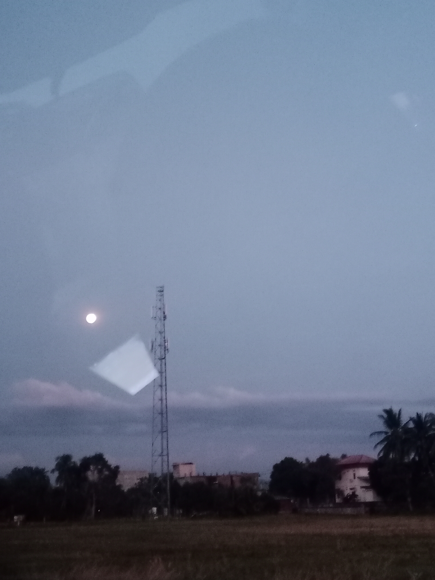 The image shows a landscape taken at dusk, likely in Cotgapu, Indonesia. In the foreground is a field of dry grass. Behind the field is a line of trees obscuring parts of buildings. A tall telecommunications tower stands to the left of the buildings. The sky is overcast, with a dim moon visible. There's a bright square glare, likely a reflection on a window from which the photo was taken. Palm trees are visible on the right of the image.