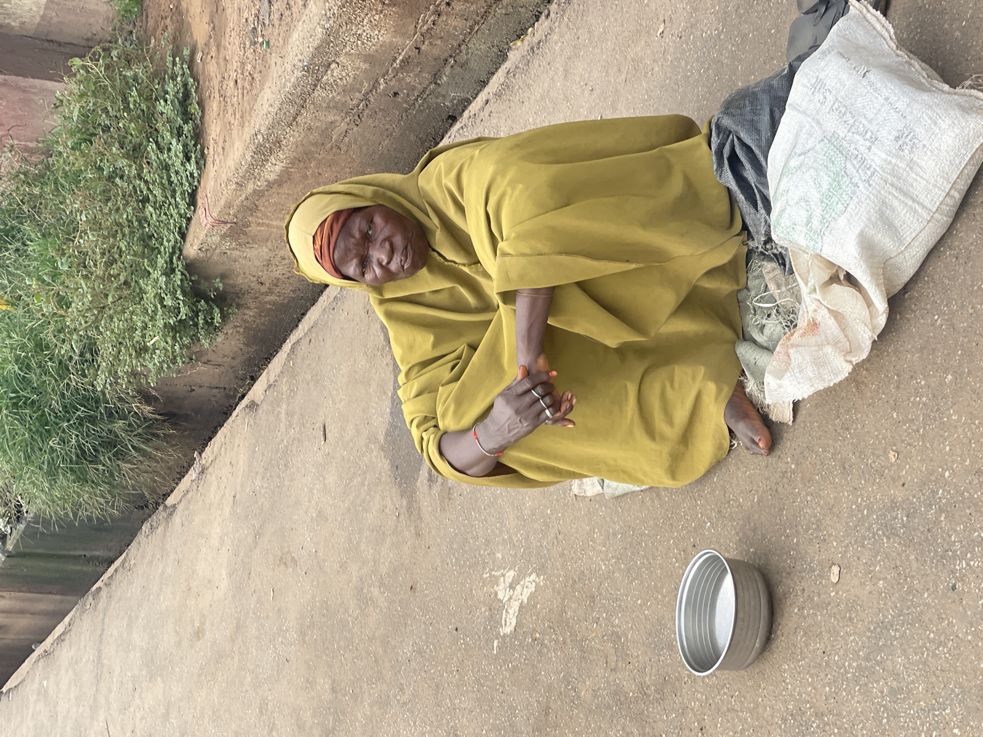 An elderly woman, dressed in a yellow headscarf and matching cloak, sits on a concrete surface, her legs crossed. In front of her, a metal bowl rests on the ground. Behind her are several bags, one white with green writing, and a dark gray one. The woman has her hands clasped in her lap. The background shows a concrete drainage ditch with lush green vegetation growing from it. The scene is set on a road in Lokoja, Nigeria.