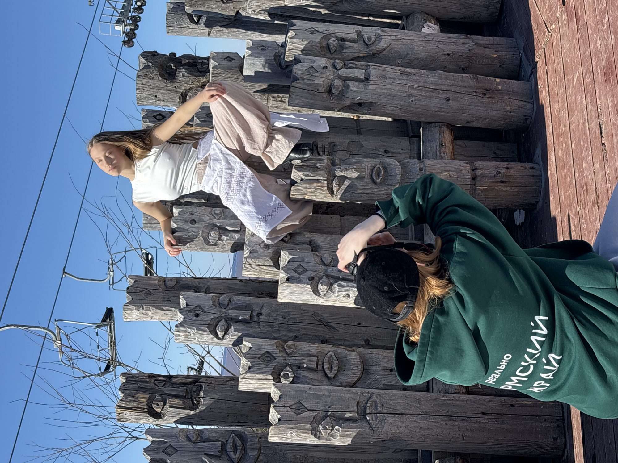 A photo taken in Gubakha, Russia shows a young female, with light brown hair, wearing a white t-shirt and long white and beige skirt, posing on a structure made of carved wooden logs, while another person in a green hoodie and black baseball cap kneels to photograph her. The backdrop features a clear blue sky and a ski lift structure in the distance. The carved logs have repetitive facial designs, adding a cultural or artistic dimension to the scene. A wooden platform is partially visible at the right edge.