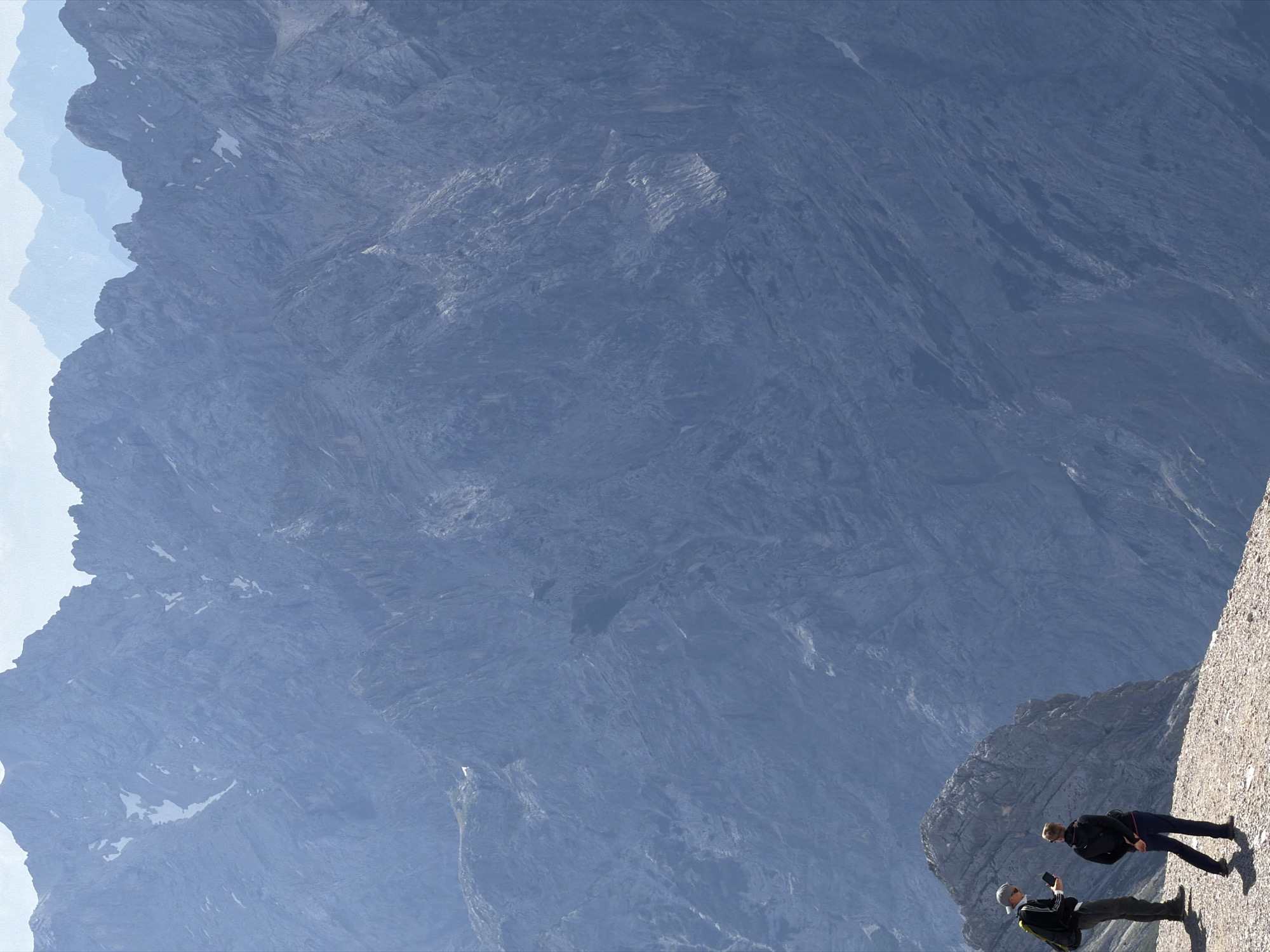 The image depicts a mountainous landscape with two individuals standing on a rocky ridge, looking out at the vast expanse. The foremost individual, wearing a grey cap, dark jacket with white stripes, and dark pants, is holding a smartphone and what appears to be a portable device. The second individual, facing away from the camera and wearing a black jacket and dark pants, has their hands clasped behind their back. They are positioned on a gravelly, uneven surface, with a large, craggy rock formation to their left.
In the background, a massive, blue-hued mountain range stretches into the distance, its peaks fading into a hazy sky. The mountains are characterized by steep, rugged slopes and visible rock strata. Patches of snow are visible on some of the higher elevations, suggesting a high altitude. The overall atmosphere is one of grandeur and isolation, with the scale of the mountains dwarting the human figures.
This scene is likely located in the vicinity of Garmisch-Partenkirchen, Germany, a region renowned for its dramatic alpine scenery and popular hiking destinations. The rugged terrain and the presence of hikers are consistent with the natural environment found in the Bavarian Alps, where Garmisch-Partenkirchen is situated. The image captures a moment of observation and appreciation of the natural world from a high vantage point.