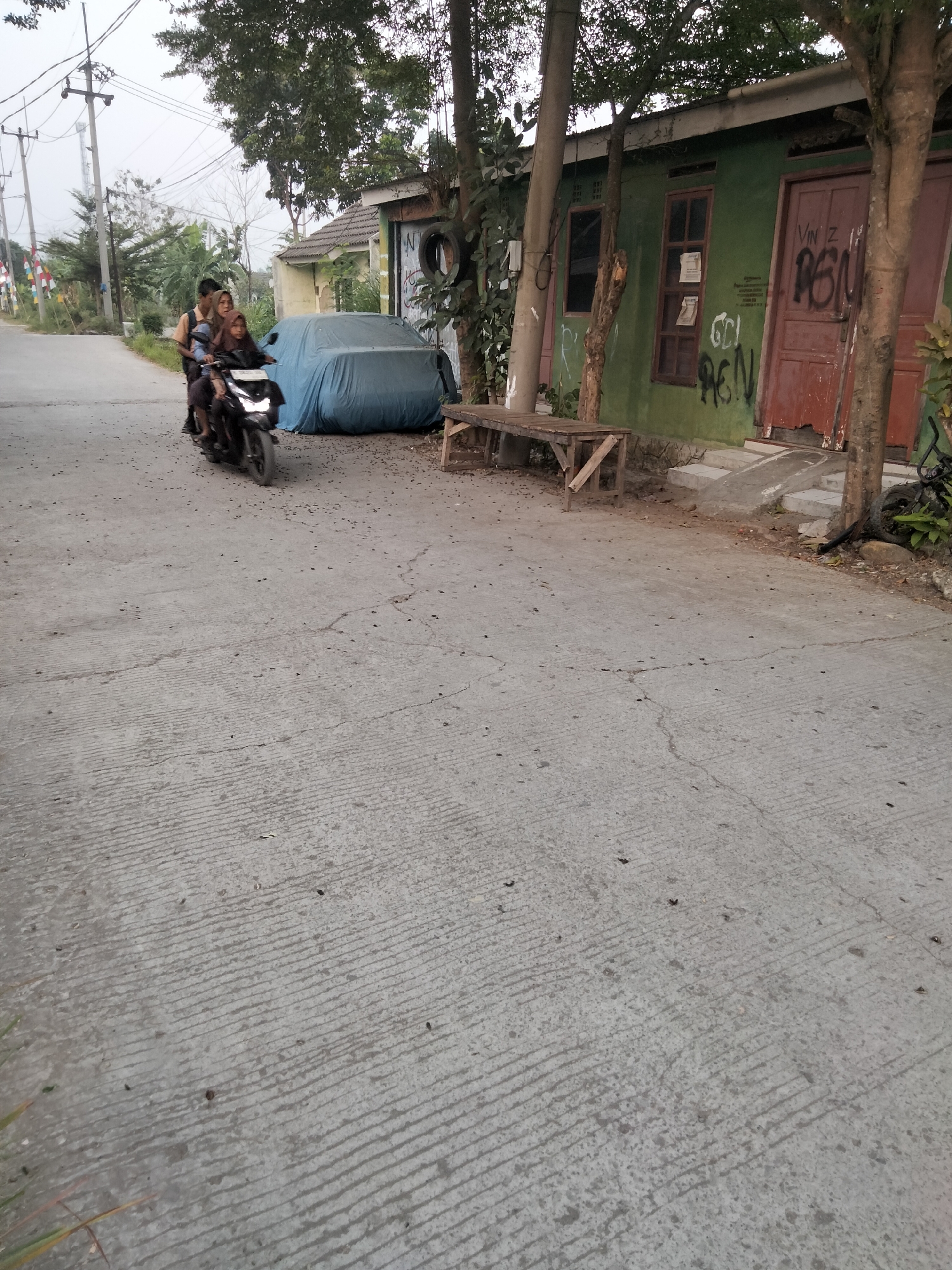 In Cariu, Indonesia, a motorcycle carrying three individuals rides on a paved road. The motorcycle is black and the riders, two children and one adult, are seated behind each other. The child in the front is wearing a brown shirt, the middle child is wearing a blue shirt, and the adult in the back is wearing a dark blue shirt. The road is cracked and textured, with scattered debris. To the right of the road, a single-story green building with a brown door and windows is visible. Graffiti is spray-painted on the building's facade. Adjacent to the building is a wooden bench and a car covered with a blue tarp. Trees with dense foliage are present on both sides of the scene, and utility poles with wires stand to the left. The sky is overcast.