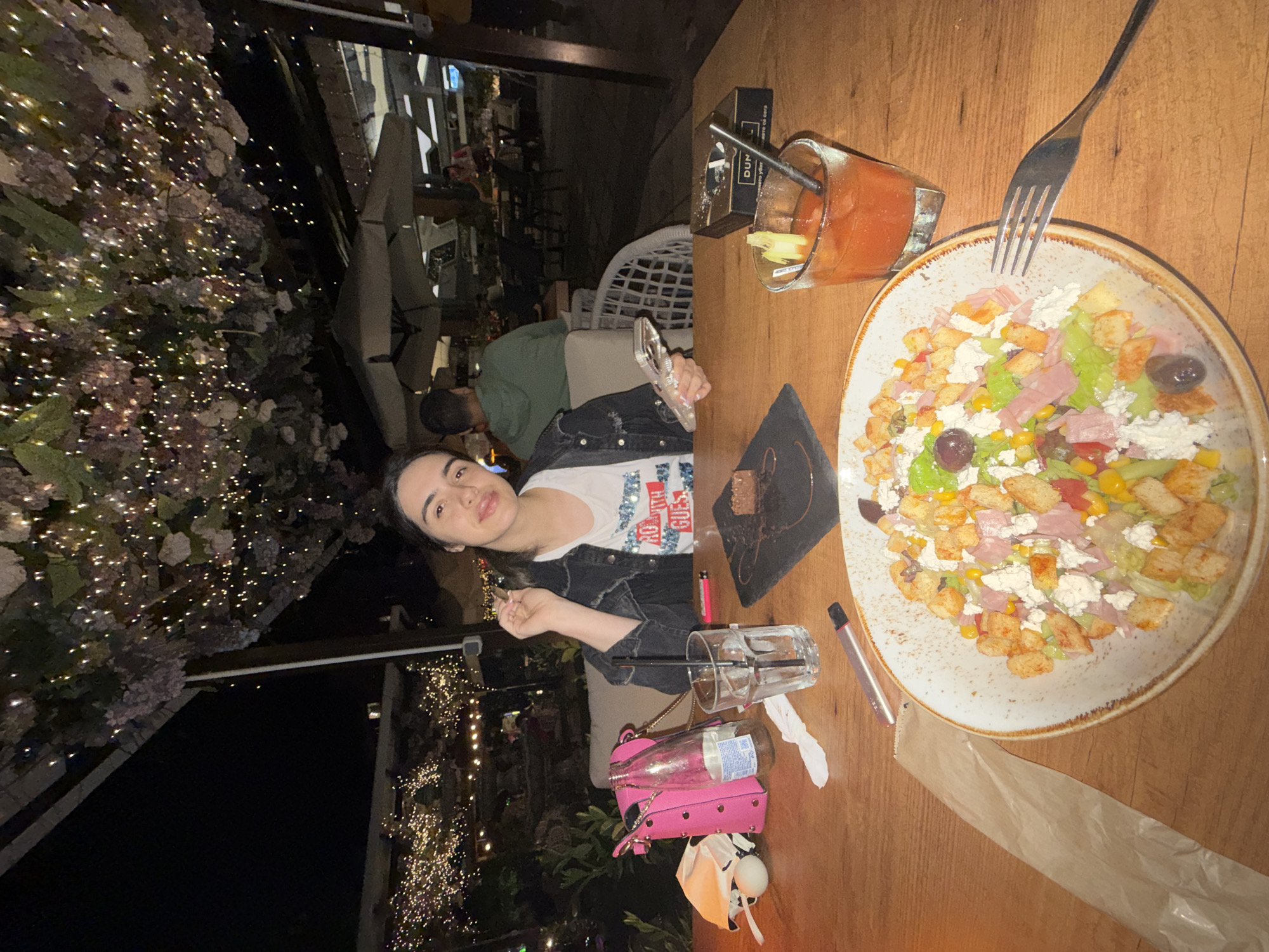 The image shows a woman at a wooden table, presumably at a restaurant or cafe, possibly in Sofia, Bulgaria. The setting appears to be outdoors at night, as evidenced by the festive lights and the presence of outdoor seating arrangements in the background.
The woman is seated at the table, which is cluttered with various items. There's a plate of salad, a drink with a straw, what appears to be a small dessert on a slate board, a lighter, an electronic cigarette device, an empty glass, and a pink purse. A fork is poised over the salad plate.
The woman appears to be holding something small in her hand. Another person is partially visible behind her.