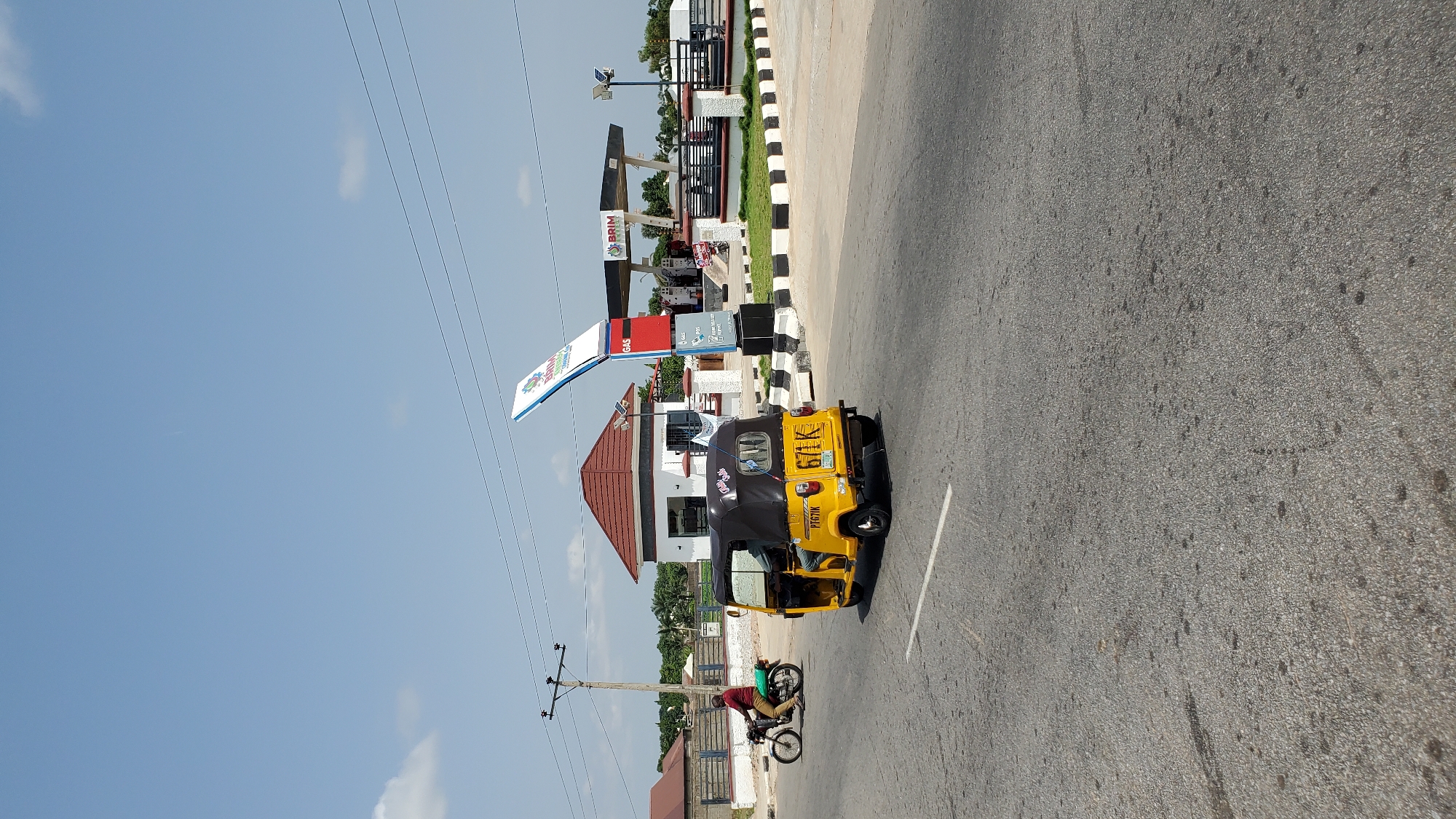The image shows a street scene in Jalingo, Nigeria, featuring a yellow auto-rickshaw driving on the right side of the road. In the background, there is a gas station with a prominent sign. A person is riding a motorcycle on the left side of the road, heading in the opposite direction of the auto-rickshaw. The sky is clear with a few scattered clouds. The road surface appears to be asphalt, and there is a white line marking the lane. The environment suggests a typical urban or semi-urban setting in Nigeria.