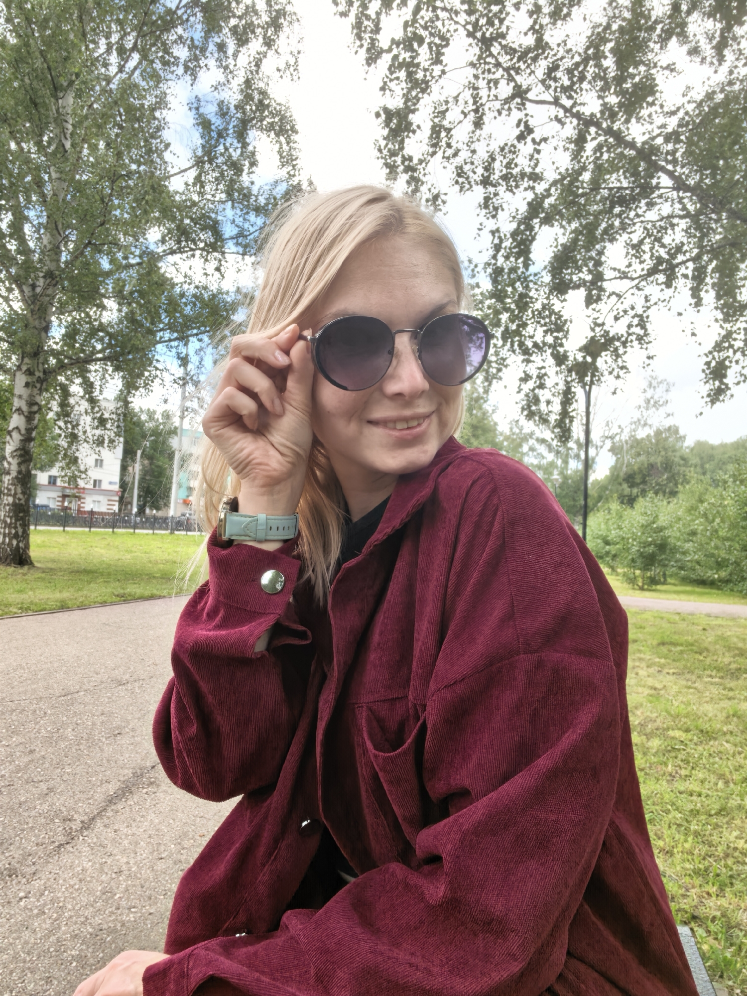 The image shows a young blonde woman wearing sunglasses and a maroon corduroy jacket. She is sitting outdoors, likely in a park or public space, as indicated by the presence of trees, grass, and a paved walkway. In the background, there are buildings visible, suggesting an urban setting. The woman is adjusting her sunglasses with her right hand. Her left wrist has a watch with a light blue strap. The overall scene is bright and appears to be taken during the daytime. The specific location in the city of Sterlitamak, Russia, cannot be determined from the visual information provided in the image.