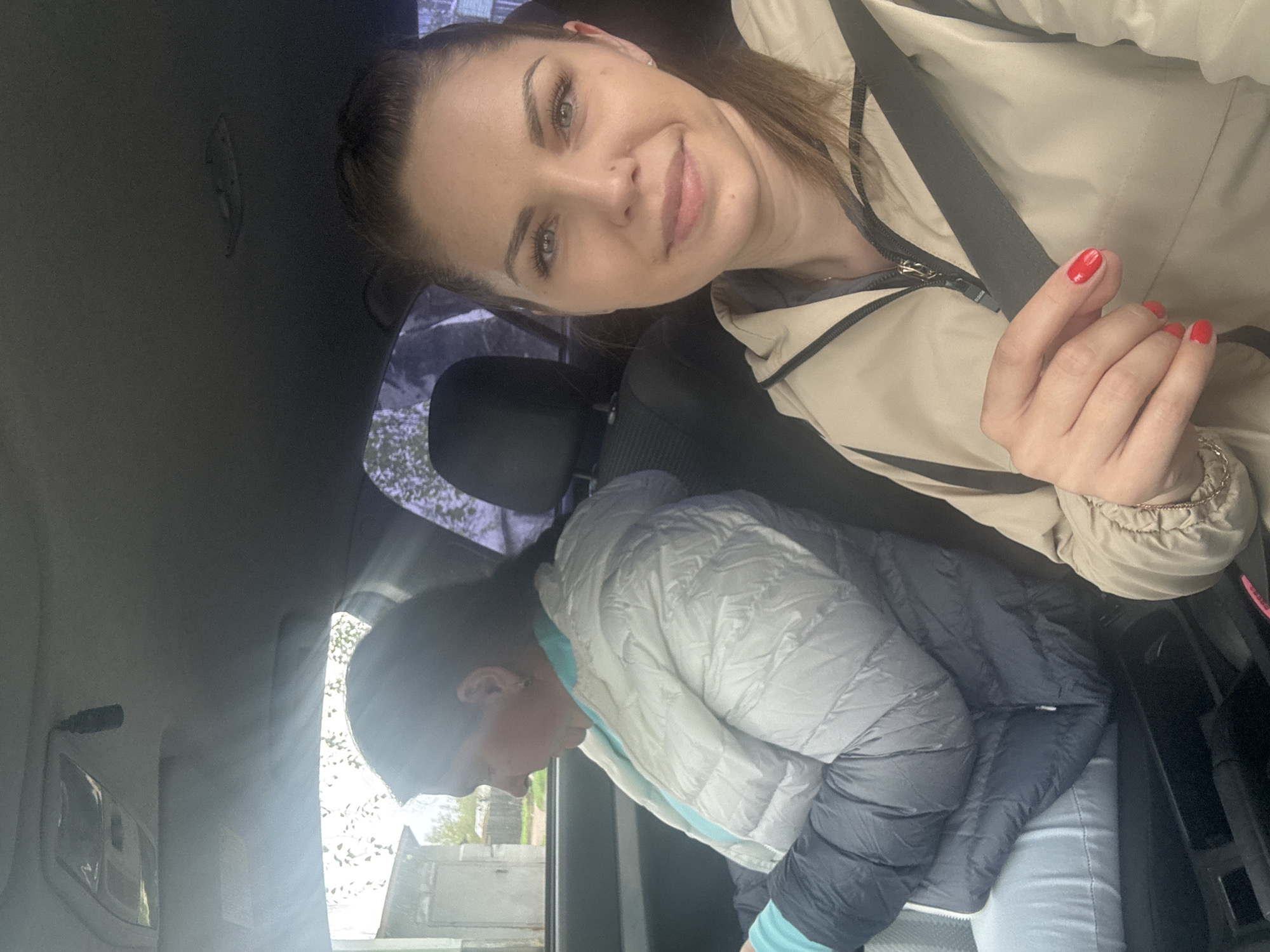Two women are in a car. One woman, in a beige jacket with red nails, is taking a selfie. She is in the passenger seat and wearing a seatbelt. The second woman is in the back seat, wearing a white and grey puffer jacket with a light blue undershirt. Outside the car window, a building is visible. The location is Раздолье, Russia.