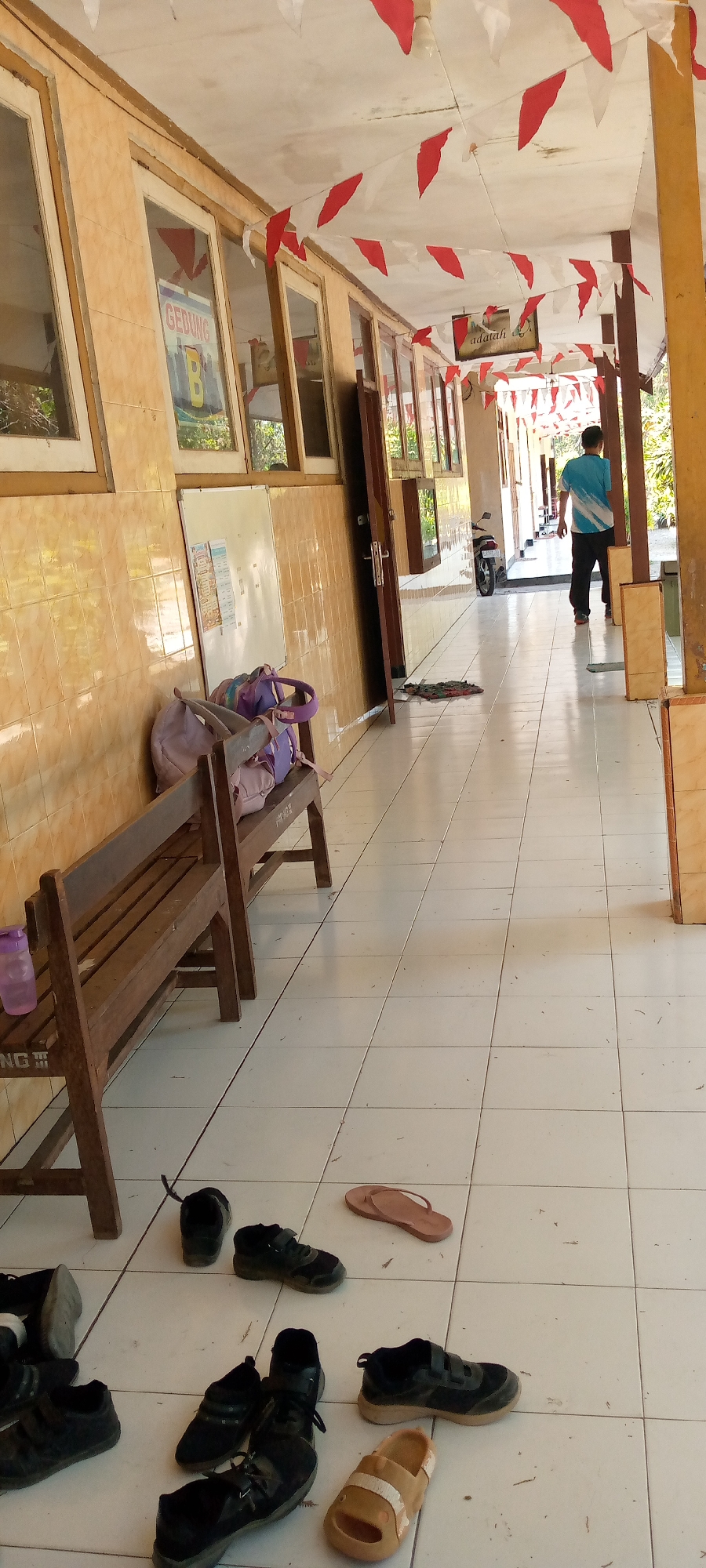 The image depicts a hallway at a school in Pacing, Indonesia, decorated with red and white triangular flags. In the foreground, several pairs of shoes and one pair of flip-flops are scattered on the tiled floor. A wooden bench with two purple bags on it sits to the left, and a water bottle is on the bench. Further down the hallway, a person in a blue and white shirt walks away from the camera. A motorcycle is parked on the right side of the hallway. The walls are tiled with a yellowish-brown pattern, and several windows with wooden frames are visible along the wall. Some posters are displayed on the wall, including one that reads "GEDUNG B" (Building B). The floor is made of white square tiles. The lighting suggests it is daytime, with sunlight streaming in from the right side.