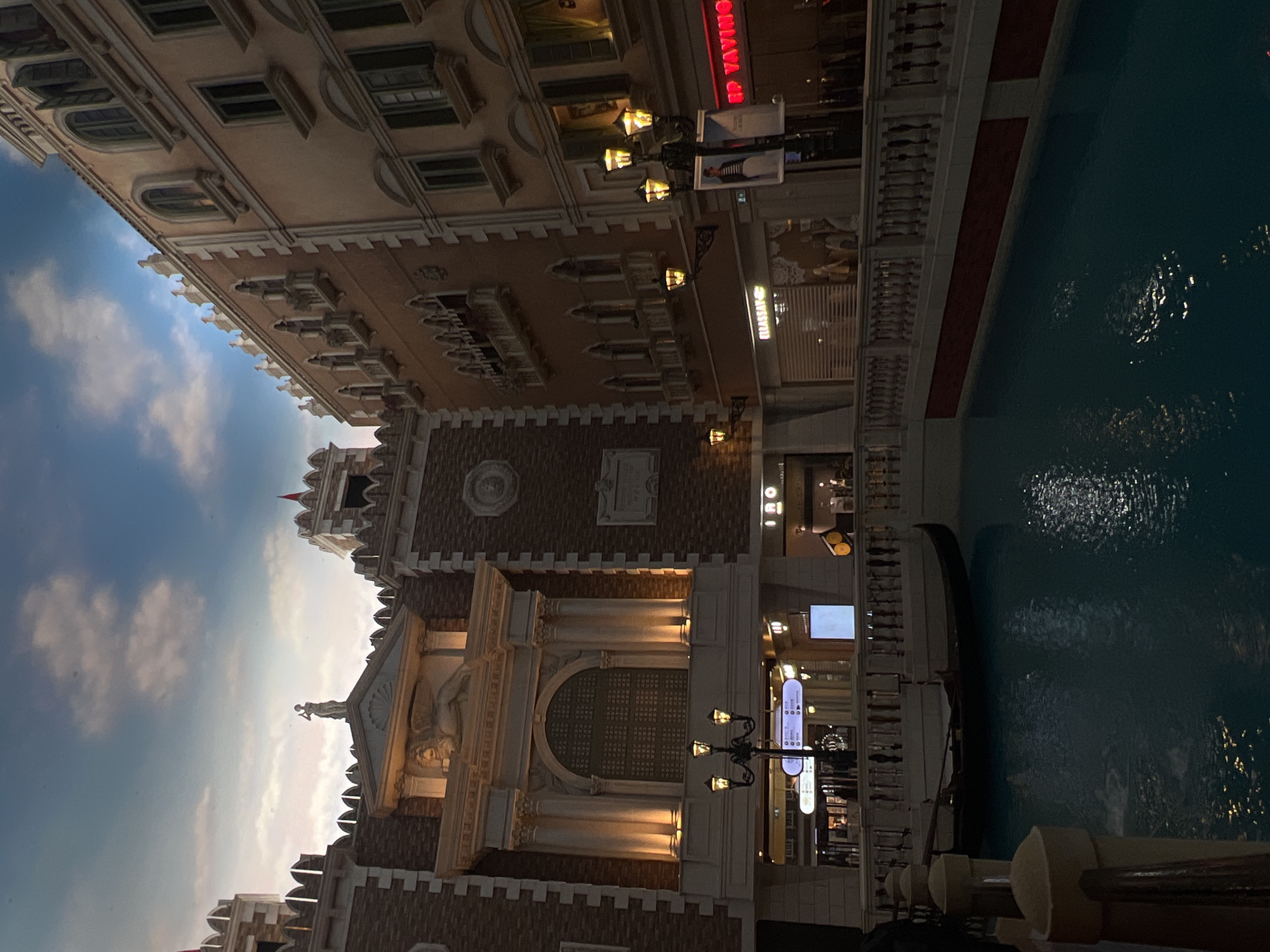 The image shows a view of a Venetian-style building complex, likely a hotel or resort. The architecture is reminiscent of Venice, with canals running alongside the buildings. There's a gondola in the water, indicating a leisure or touristy environment. The sky is partly cloudy. A sign "EP YAVIMOS" is visible in the upper right. The location is likely a tourist attraction in Unknown, Macao, given the theme and architecture. A statue can be seen on top of the building. There are lampposts adding to the Venetian atmosphere.