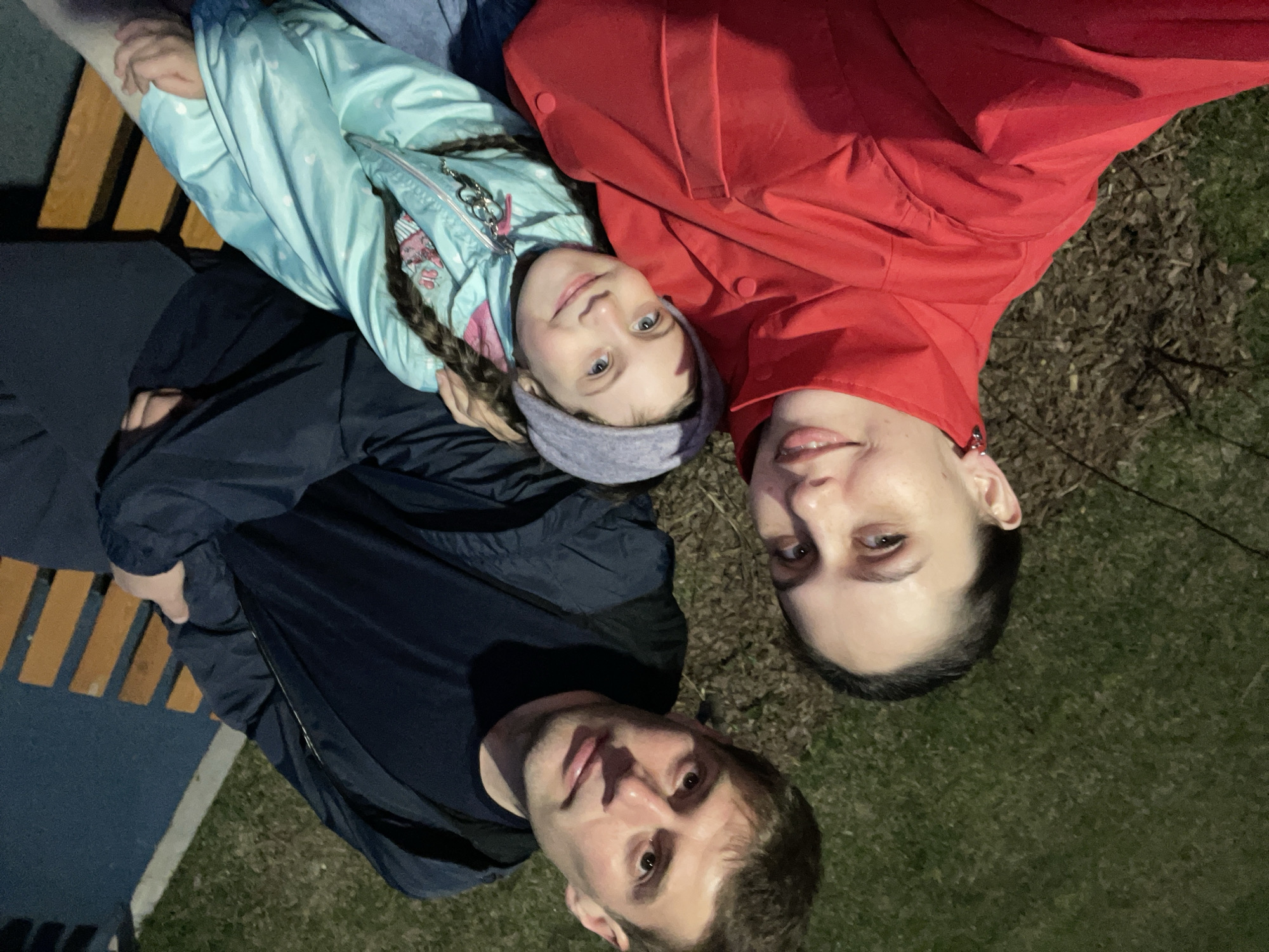 A family of three is posing for a selfie outdoors at night. A man wearing a dark jacket is at the bottom of the frame. Above him is a woman in a red jacket. To her left is a girl with braided hair, wearing a light blue jacket and a headband. They are likely sitting or lying on the grass, which is visible in the upper right corner. A park bench and a paved area are visible in the lower left corner. The image does not provide enough information to determine the precise location in Izhevsk.