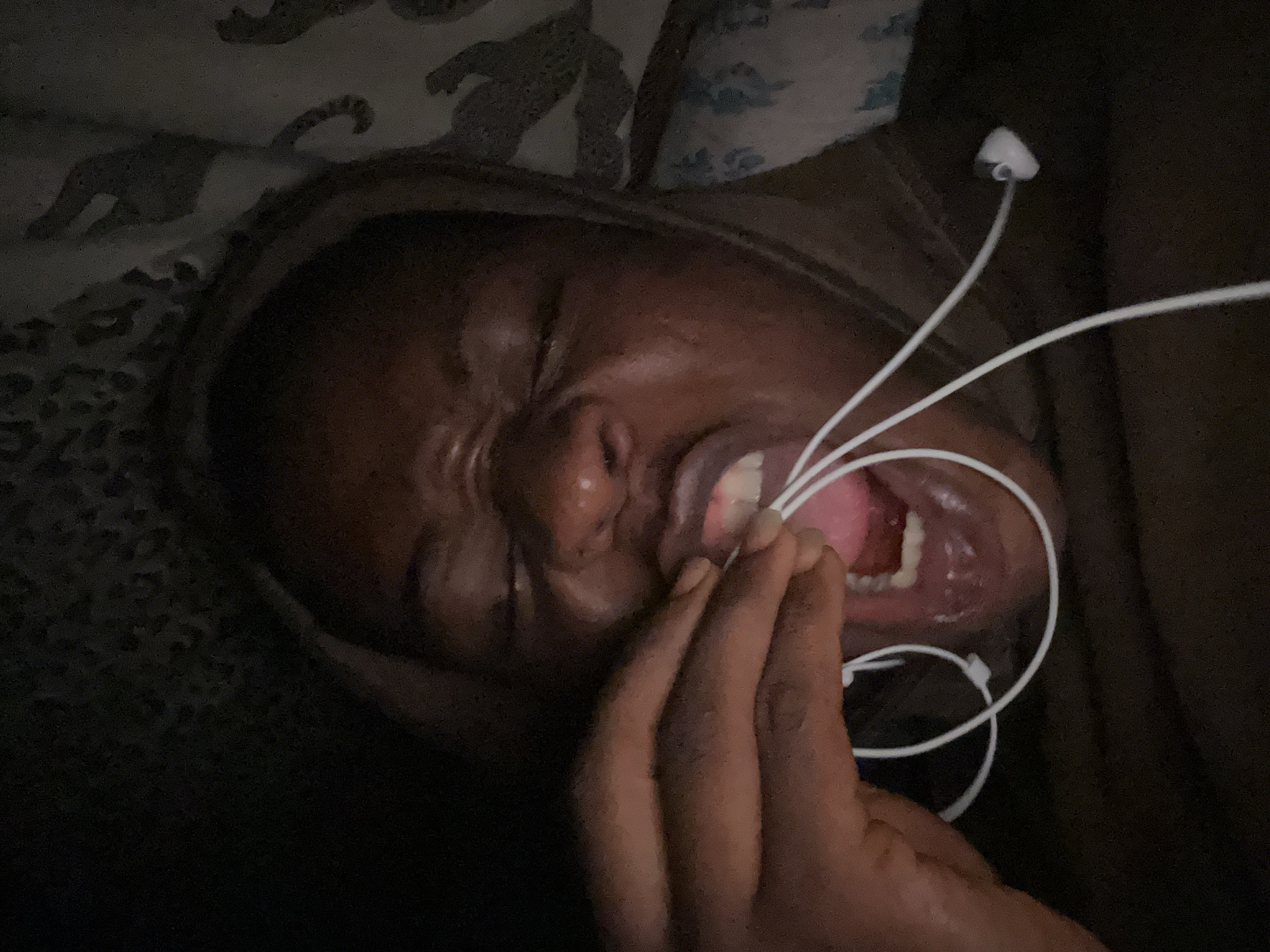 A close-up, low-light, and angled image depicts a person with dark skin in an indoor setting. The person is lying down, possibly in a bed or similar resting place, as suggested by the patterned bedding partially visible in the upper left corner. The person's eyes are closed, and their mouth is open, with visible teeth and tongue. A hand is positioned near the mouth, with the fingers appearing to touch or hold white earphone cords. The earphone cords are tangled and extend around the person's face. The overall environment is dark and shadowed, obscuring specific details of the room or location. The city and location are unknown.