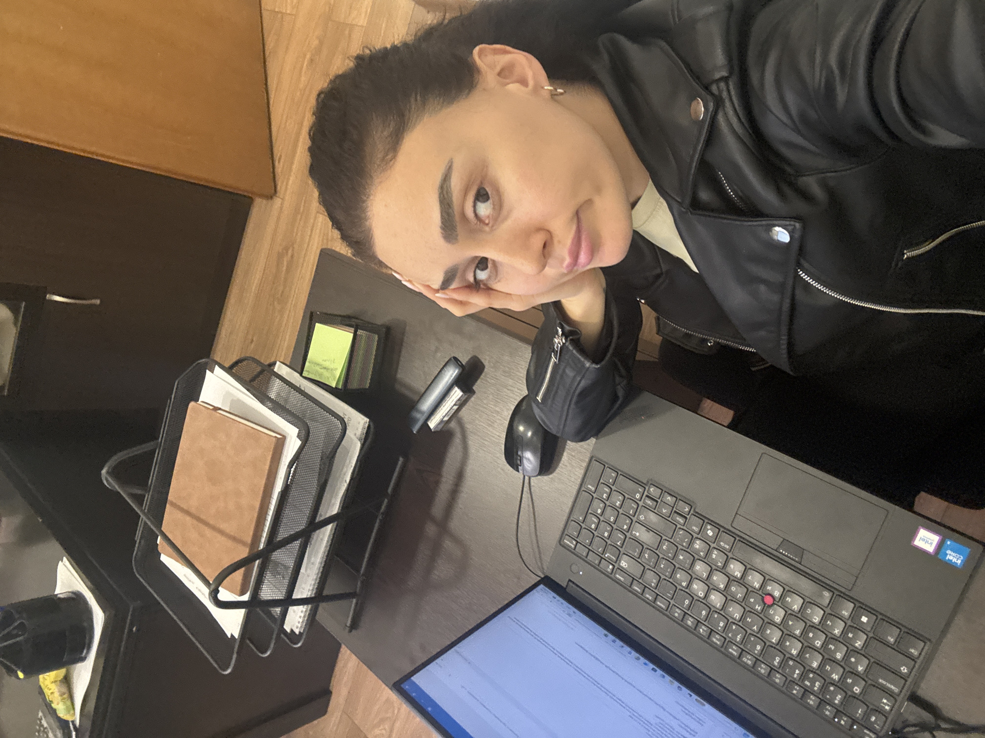 A woman with dark hair pulled back is photographed at an angled, overhead perspective while resting her chin on her hand. She is wearing a black leather jacket. In front of her is a dark-colored desk with a laptop displaying text, a mouse, a stapler, a stack of sticky notes, and a black wire file organizer filled with a notebook and papers. A cabinet with a wood-tone door is partially visible in the upper left corner. The location cannot be determined from the image.