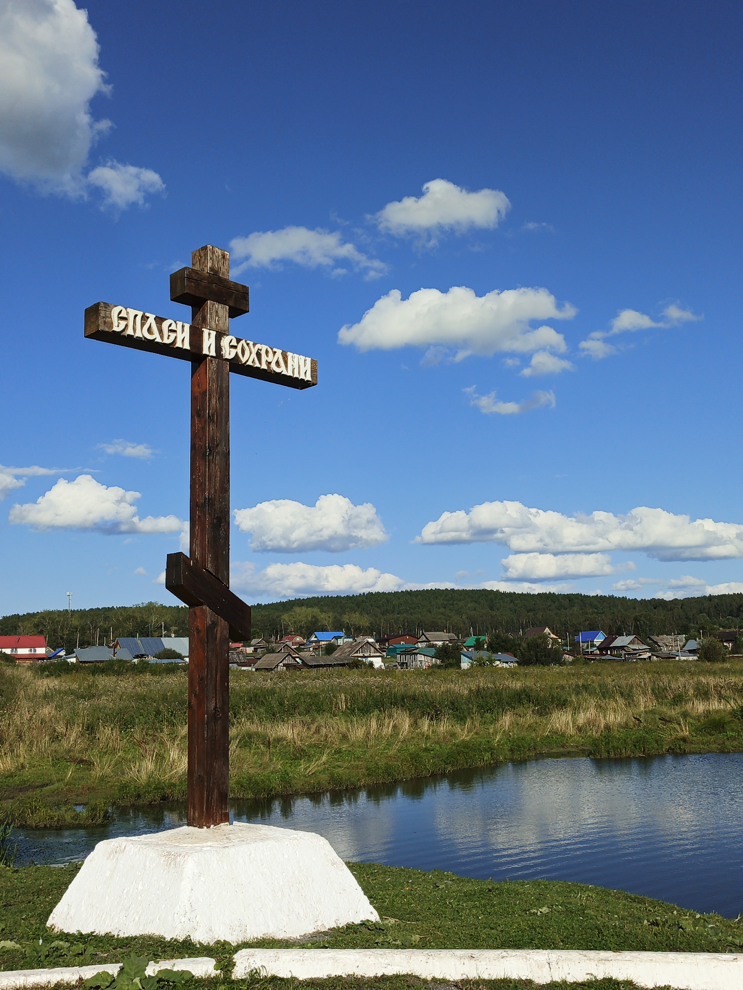 A wooden Orthodox cross stands in the foreground on a white pedestal. The cross is dark brown with white Cyrillic lettering that reads "СПАСИ И СОХРАНИ" (Save and Protect). The cross is oriented vertically, with a smaller horizontal beam near the top and a slanted beam below it. Behind the cross, a body of water reflects the blue sky and clouds. The background consists of a grassy field, a village with various houses with colorful roofs, and a dense forest on a hill. The sky is a bright blue with scattered white cumulus clouds. The scene appears to be in the village of Maly Berdyaush, Russia, as indicated by the distinctive architecture and the presence of an Orthodox cross. There are no people visible in the image.