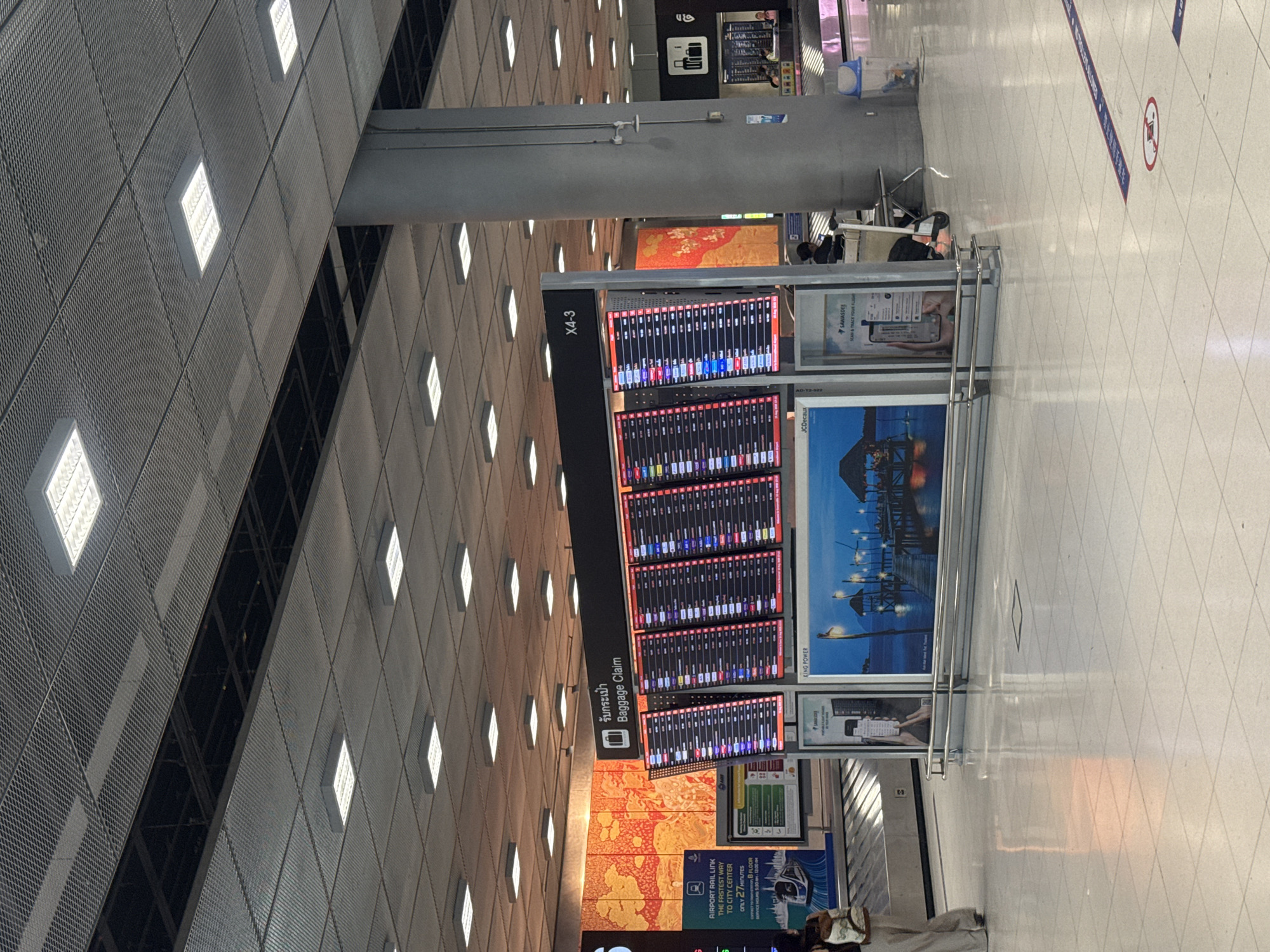 An indoor airport baggage claim area is depicted in this vertical, eye-level, medium-shot image. The scene is brightly lit by fluorescent ceiling lights and features a large digital departures board displaying flight information. The board is mounted on a dark frame with a sign that reads "Baggage Claim" in English and Thai script. To the left of the departures board is a colorful mural with abstract orange and yellow patterns. In front of the departures board, there is a baggage carousel with luggage. Two individuals are visible walking away from the carousel in the lower left corner of the frame. To the right of the departures board, a large cylindrical pillar supports the ceiling. Further to the right, a trash can and a few scattered items are visible on the floor. The floor is tiled in a light color, and a blue and white social distancing marker is present on the floor in the lower right portion of the image. The background shows more of the airport environment, including a distant baggage carousel and more digital signage. The image appears to be taken in Nong Prue Subdistrict, Thailand, given the Thai script on the signage.