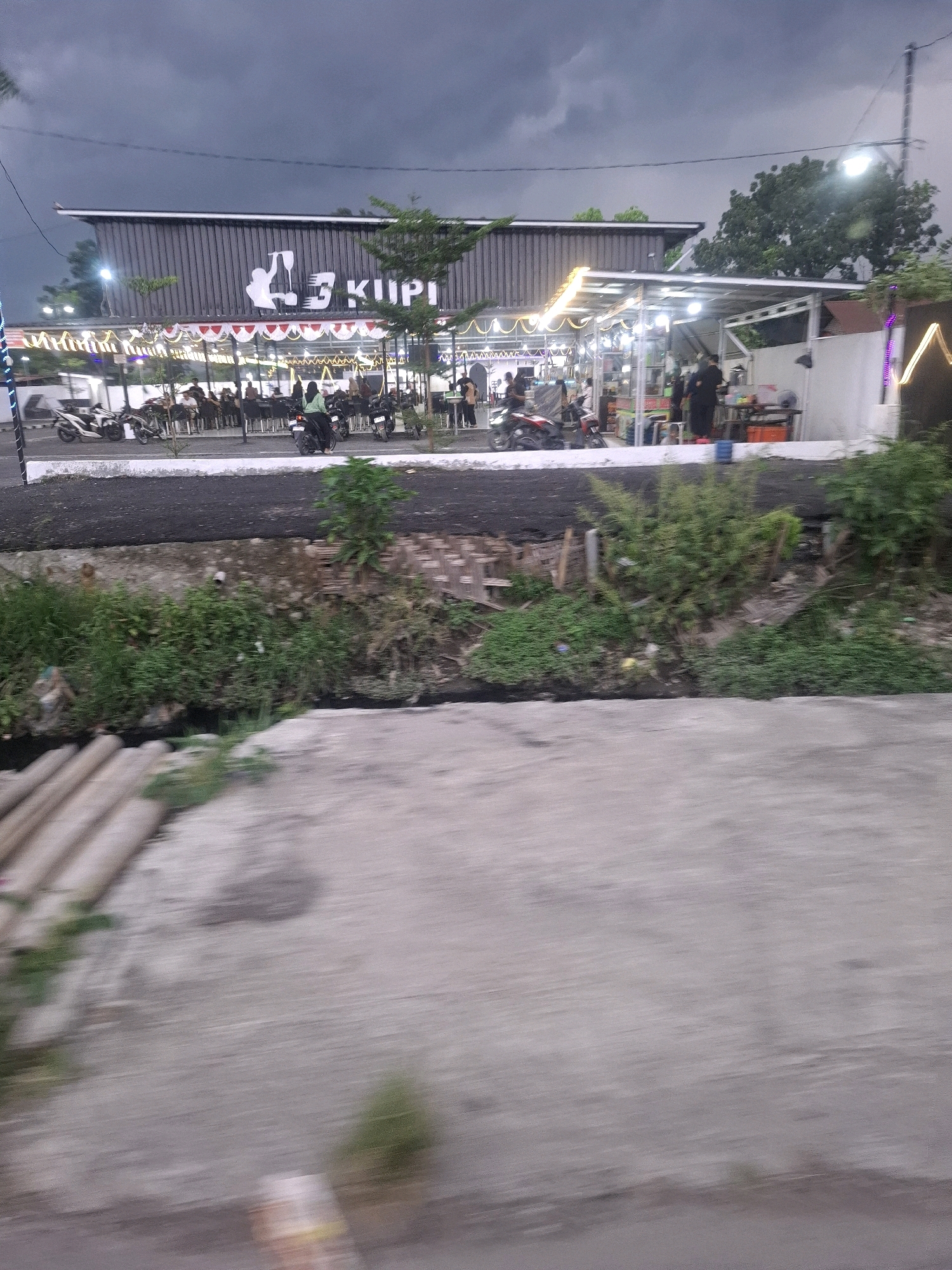 The image shows a street-level view of a "Kopi" establishment in Kenangan, Indonesia. The building features a dark corrugated metal facade with a white logo depicting a coffee cup and the text "3 KUPI". In front of the building, there are several tables and chairs set up under a covered outdoor seating area adorned with strings of lights. People are seated at some of the tables. Several motorcycles are parked in front of the establishment, and some individuals are interacting with them. There are food stalls or kiosks set up further down the street, also lit by strings of lights. The environment appears to be a somewhat busy urban or suburban street with a cloudy sky overhead. A ditch or canal with vegetation runs along the foreground, separating the viewer from the establishment. The foreground also shows a concrete surface, possibly a sidewalk or road.