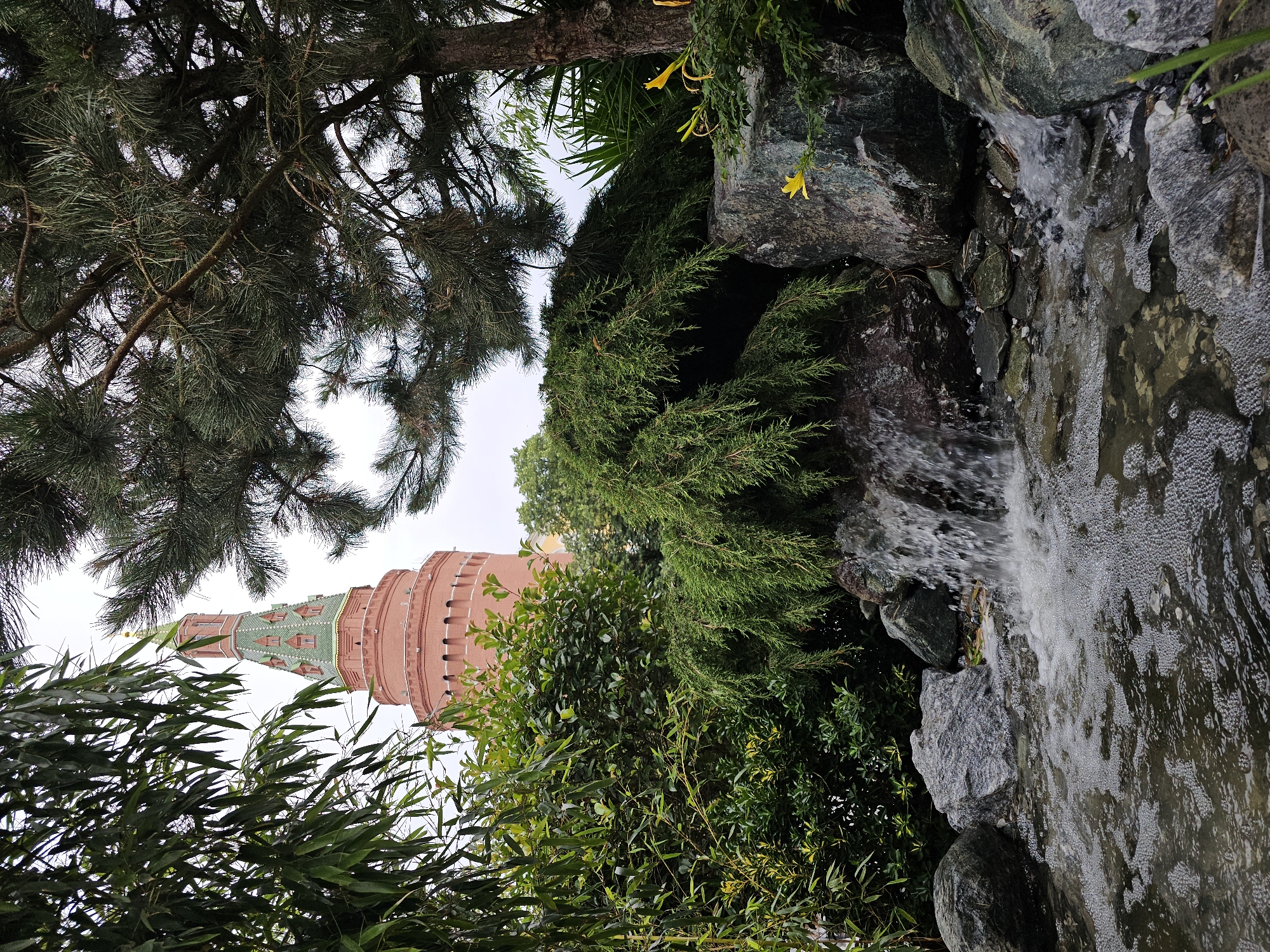 This is a vertical, eye-level shot of a small waterfall cascading over rocks into a pool of water, with foliage and a tower in the background, taken from a garden. The tower, which is made of red brick and has a green, pointed roof with gold accents, is likely a Moscow Kremlin tower. The foreground features a small waterfall with white, frothy water flowing over dark, wet rocks. The pool reflects the surrounding greenery and the cloudy sky. Lush green plants, including bamboo-like leaves on the left and a bushy evergreen on the right, frame the scene. In the distance, behind the waterfall and greenery, a prominent red brick tower with a green tiled roof is visible. The tower's architecture suggests it is one of the historic Kremlin towers in Moscow, Russia. The overall ambiance is serene and natural, with a striking juxtaposition of the man-made structure against the garden setting.
