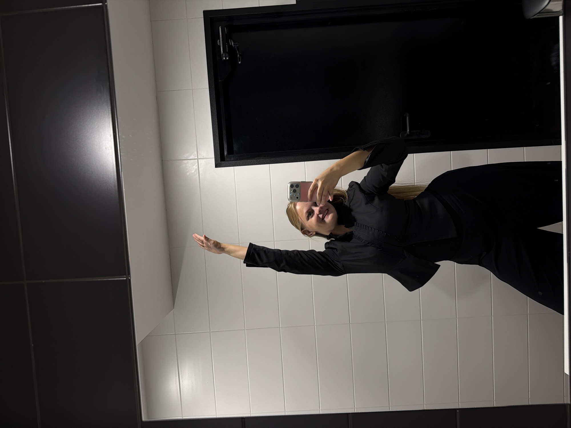 A young Caucasian woman is taking a mirror selfie in a modern bathroom. She is wearing a black blazer with ruffled details on the collar and sleeves, and black wide-leg pants. Her right arm is raised above her head, and she is smiling at her phone as she takes the picture. The bathroom features white tiled walls and a black door. The reflection shows the woman's full body and the surrounding environment. The image appears to have been taken indoors, likely in a public restroom or a similar setting. There is no information available about the location in the city of Izhevsk, Russia.