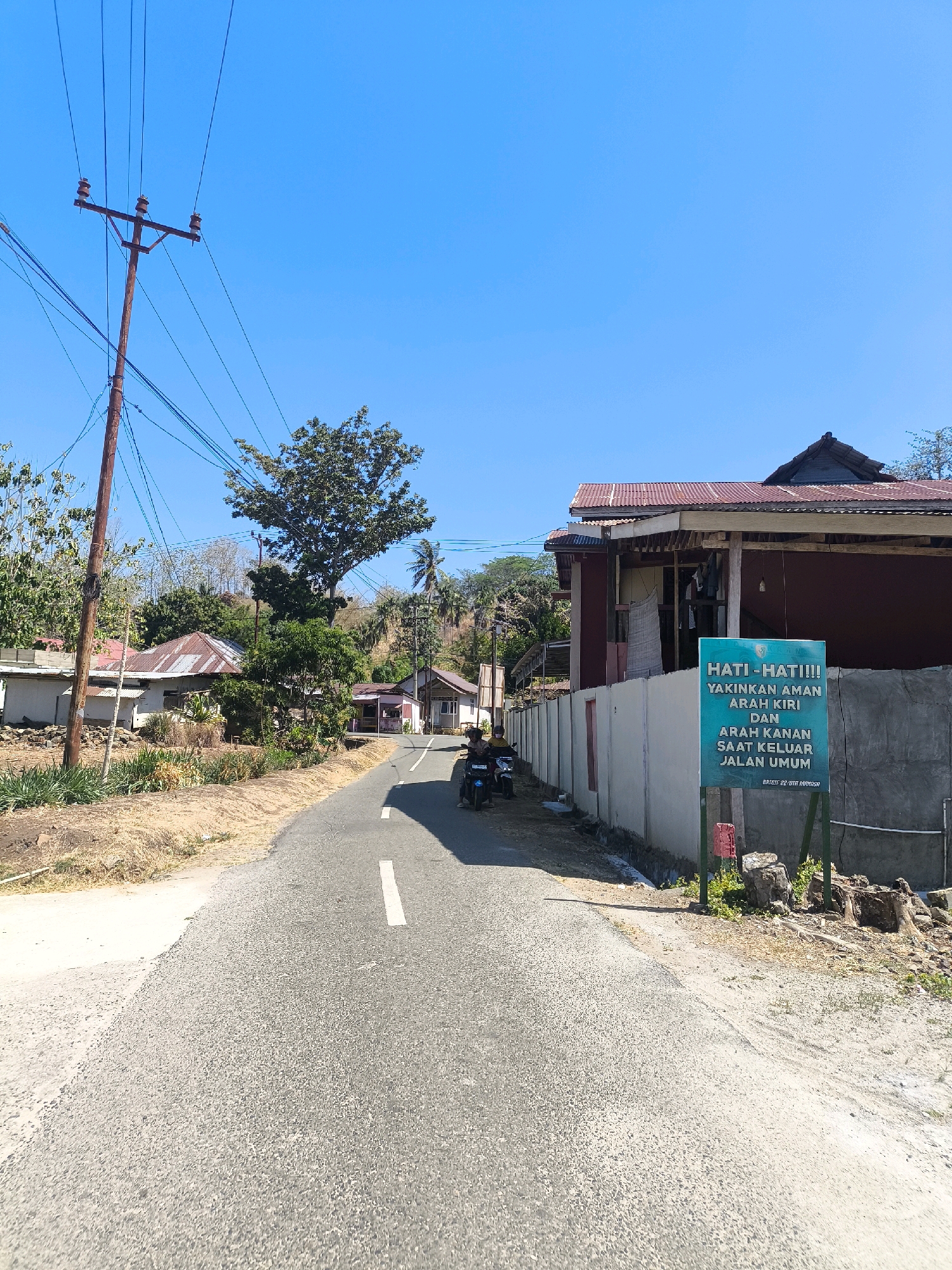 The image shows a sunny day scene on a rural road in Indonesia. A paved road with a white line down the center curves to the right in the foreground. A person on a dark-colored motorcycle is riding on the road, heading away from the camera. To the right of the road, there is a light-colored concrete wall topped with fencing, and behind it, a building with a weathered, corrugated metal roof. A green sign with text in Indonesian is mounted on a stand in front of the wall. The text on the sign reads: "HATI - HATI!!! YAKINKAN AMAN ARAH KIRI DAN ARAH KANAN SAAT KELUAR JALAN UMUM." (Be careful!!! Ensure the left and right directions are safe when exiting the public road.) The left side of the road has dry, grassy vegetation and a few scattered rocks. Further back, there are houses with similar corrugated metal roofs and trees. A utility pole with numerous wires stands on the left side of the road. The sky is clear and bright blue.