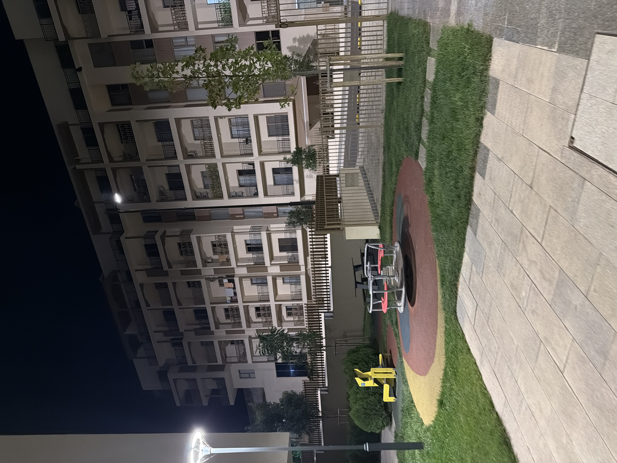 The image depicts a modern residential building at night in Chinarkent, Uzbekistan. The building features multiple stories with balconies and is illuminated by artificial light. In the foreground, a children's playground is visible, complete with a merry-go-round with red seats and a yellow climbing structure. The playground area is bordered by green grass and paved pathways. A streetlamp stands on the left side of the image, illuminating the scene. A metal fence surrounds the property, with trees and bushes planted within the grounds. The overall scene suggests a well-maintained residential complex with amenities for its residents.