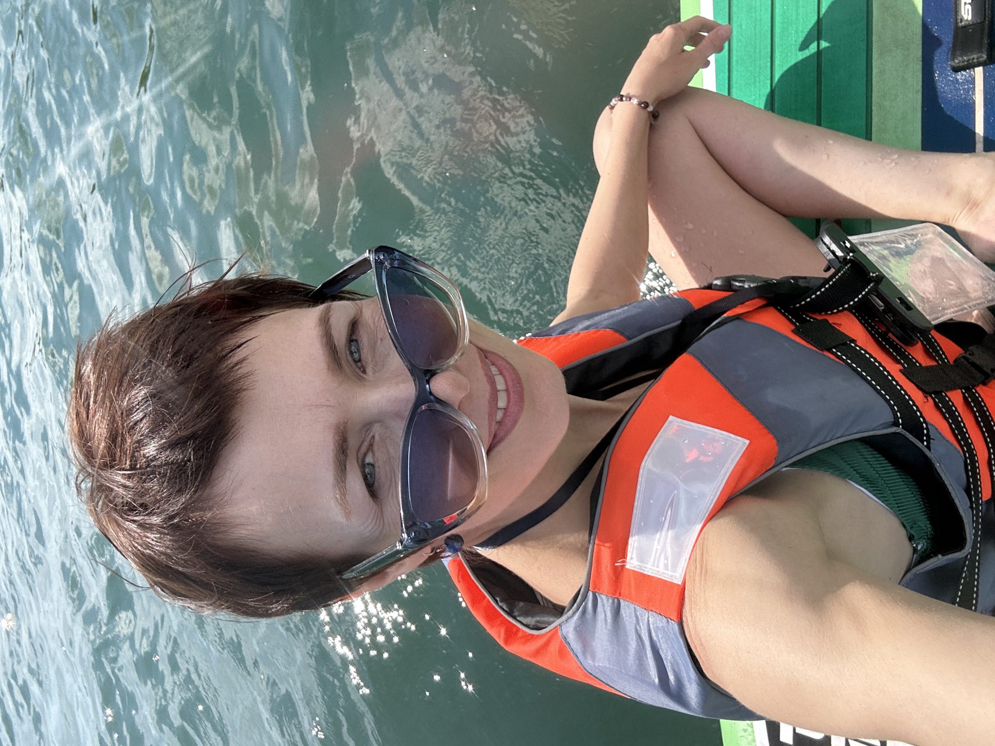 A woman with short brown hair and blue eyes is on a paddleboard. She is wearing sunglasses and an orange and grey life vest. The water is choppy and reflects the sunlight. The paddleboard is green and white. The woman is smiling. The image appears to be taken on a sunny day. The location is Kemerovo, Russia. There is text visible on the paddleboard, but it is blurry and unreadable.