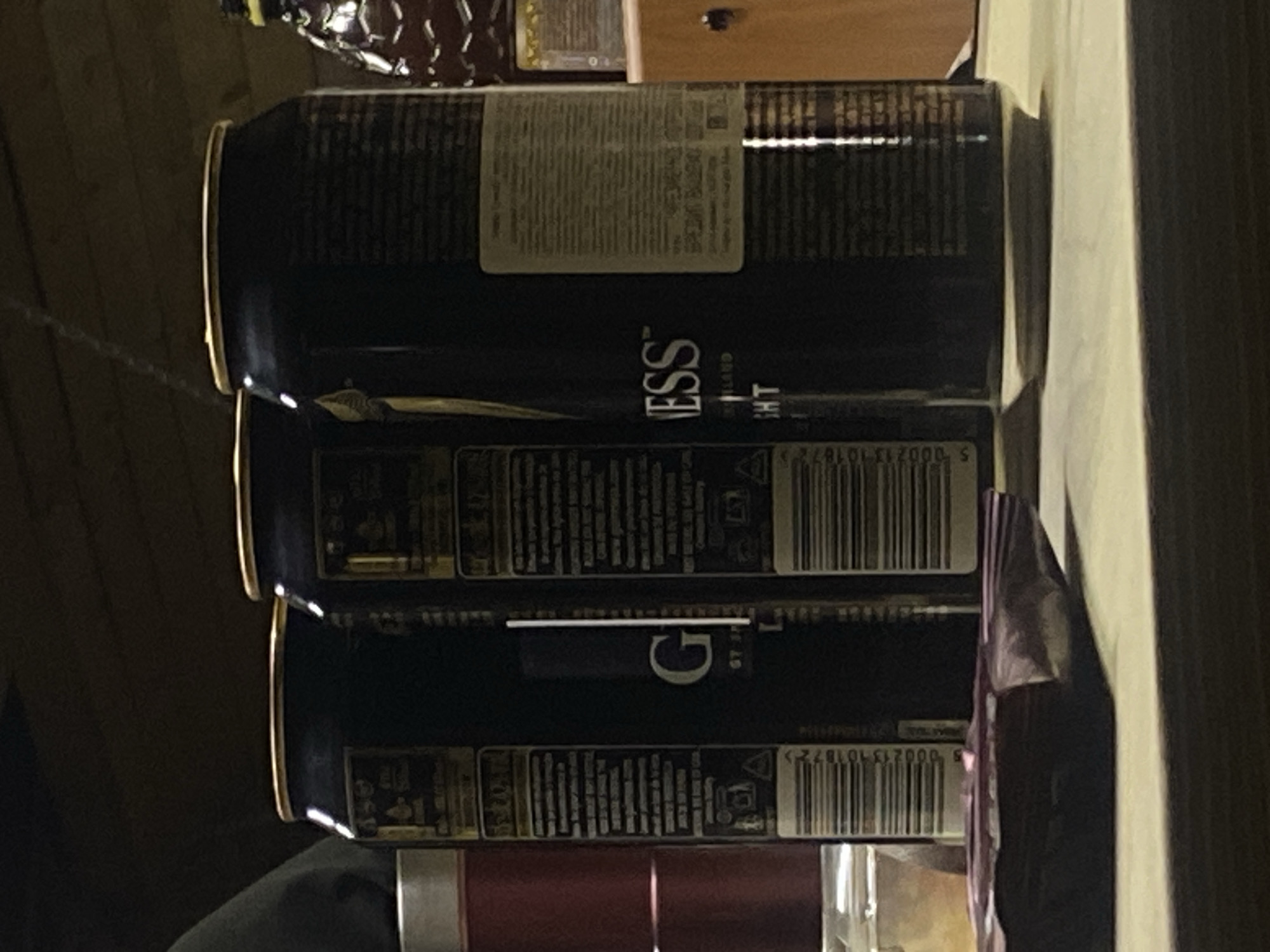 The image shows a stack of three Guinness stout cans. The cans are dark, and labels with barcodes and text are visible. The text "GUINNESS" is partially visible on the cans. The cans are stacked on a surface. Behind and above the cans, there is a bottle and a wooden surface. A reflective object is next to the can. The specific location in the city of Rusanovo, Russia is not discernible.