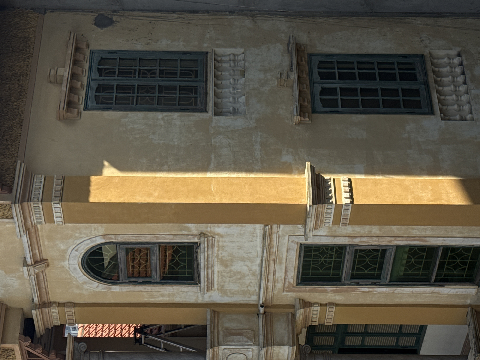 The image shows a section of an aged, yellow-toned building facade, likely located in Hà Nội, Vietnam. The architecture features decorative elements and a mix of window styles. On the upper level, there are two rectangular windows with dark frames and multi-pane glass. Between the windows are ornamental architectural details. The lower level features an arched window and a window with four sections. The lighting suggests it is daytime, and a shadow bisects the center of the image. Below the windows are structural supports and architectural embellishments. In the background, parts of other buildings and structures are visible. The overall impression is of an old building with weathered architectural character.