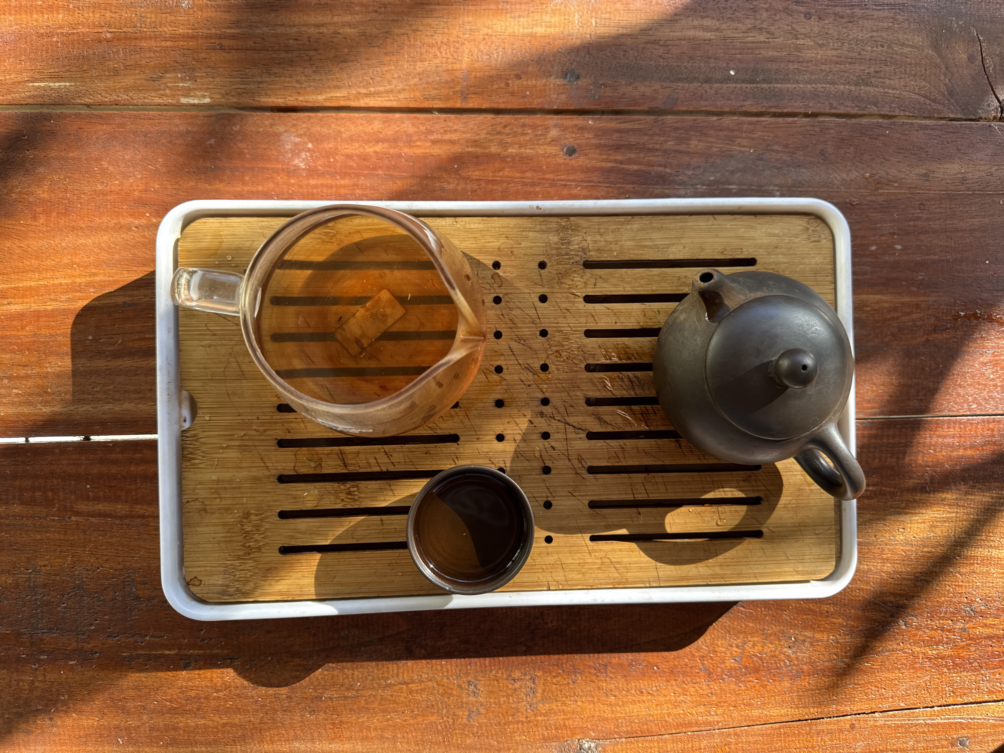 A high-angle, top-down shot captures a tea set on a wooden table in Ubud District, Indonesia. A bamboo tea tray with drainage channels rests on a wooden surface, illuminated by dappled sunlight casting long shadows. On the tray, a clear glass pitcher filled with amber-colored tea, containing ice cubes, sits to the right of a small, dark tea cup. A dark, ceramic teapot is positioned at the bottom of the tray. The scene is quiet and still, suggesting a moment of relaxation or contemplation.
