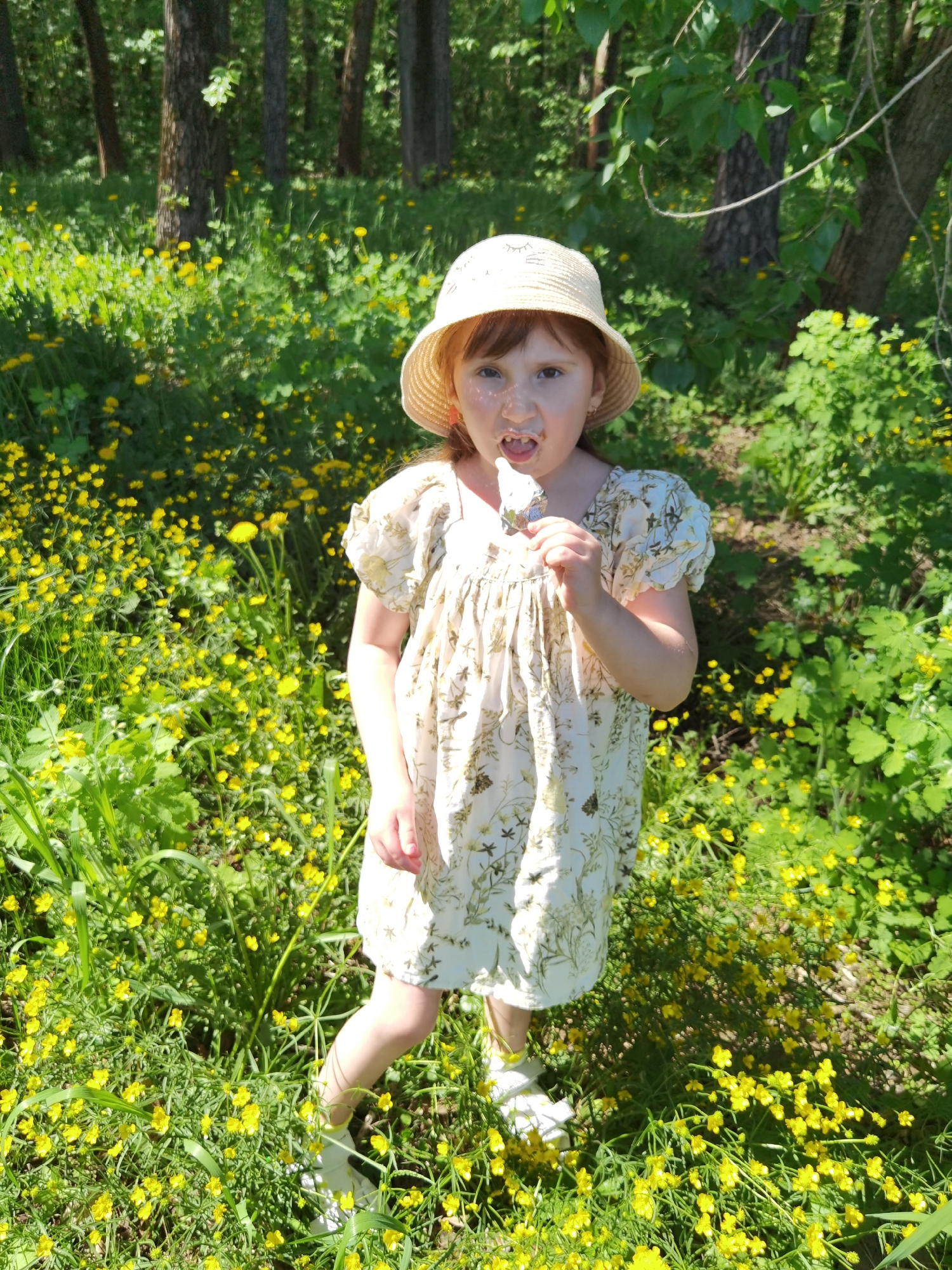 A young girl stands in a field of yellow flowers, possibly buttercups, in Perm, Russia. She wears a light-colored dress with a floral pattern, a straw sun hat, and white shoes. She is holding and eating a white ice cream or popsicle, and some of the dessert is smeared on her face. The background includes trees and green foliage, suggesting a wooded area or park. The lighting indicates a sunny day.