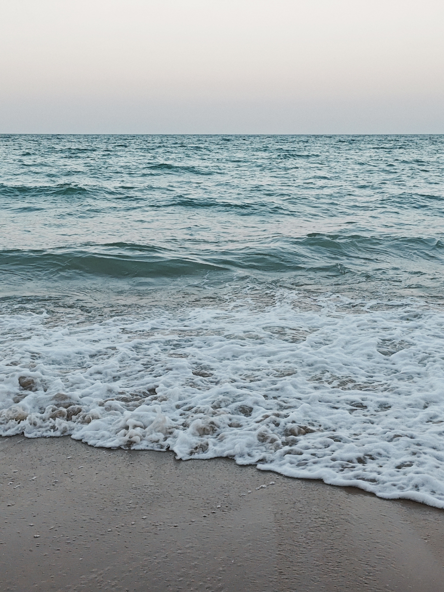 The image shows a serene beach scene with calm ocean waves gently lapping at the shore. The water is a deep turquoise color, and the sky above is a pale, muted gradient of pink and white, suggesting either dawn or dusk. The sand is a soft, light brown color, wet from the receding tide. There are no people or man-made objects visible in the frame. The composition is simple and minimalist, focusing on the natural elements of the beach and sea.