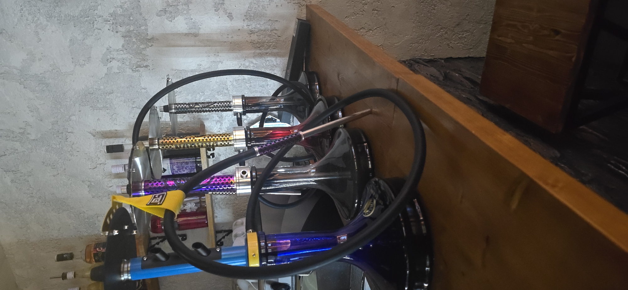 The image displays a close-up, eye-level view of several hookahs arranged on a wooden counter against a textured, light-colored wall. Four hookahs are prominently featured, with their bases in shades of blue, black, and clear glass. The stems of the hookahs exhibit intricate designs in purple, gold, and black. Black hoses are attached to each hookah, curving in front of the main subjects.
In the background, shelves hold various bottles, some appearing to be alcohol or syrups, and other glassware. The wall has a rough, stucco-like texture. The wooden counter appears to be made of rough-hewn wood with a visible grain and a reddish-brown stain. A portion of a dark wooden stool is visible in the lower right corner. The lighting suggests an indoor environment with ambient light.
No people are visible in the image. The scene appears to be a retail or hospitality setting, possibly a bar or a lounge, and the hookahs suggest a place where shisha smoking is offered. The city of Krasnodar, Russia, is not depicted or implied in the visual elements of the image.