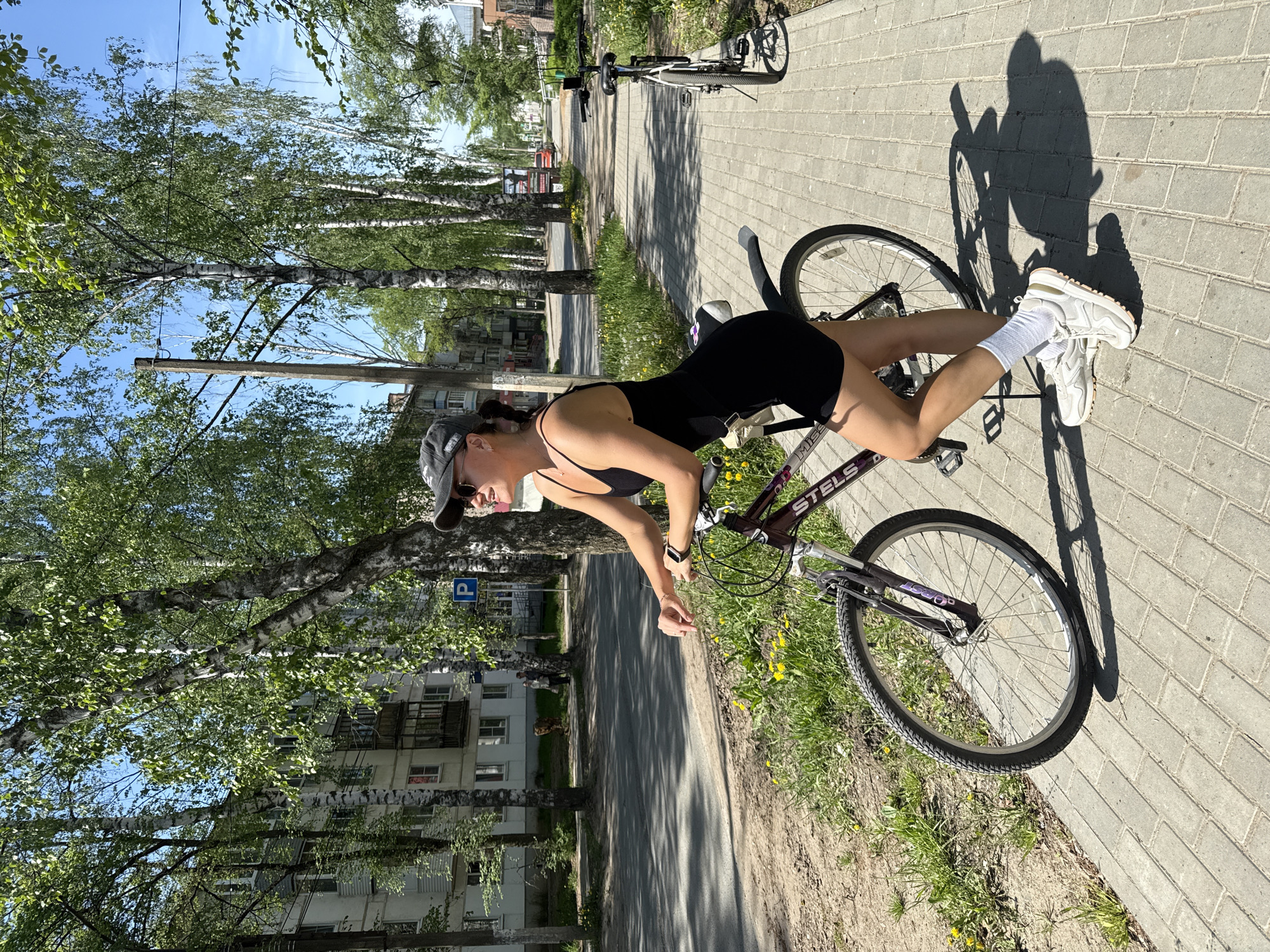 A young woman is posed on a bicycle on a brick-paved sidewalk in Chaikovsky, Russia. She is wearing a black leotard, white socks, and white sneakers, and a baseball cap. The bicycle is a dark-colored Stels mountain bike. Behind the woman and the bicycle, there is a street with birch trees lining the side and residential buildings visible. A parking sign is visible among the trees. The weather appears to be sunny. Another bicycle is parked farther down the sidewalk.