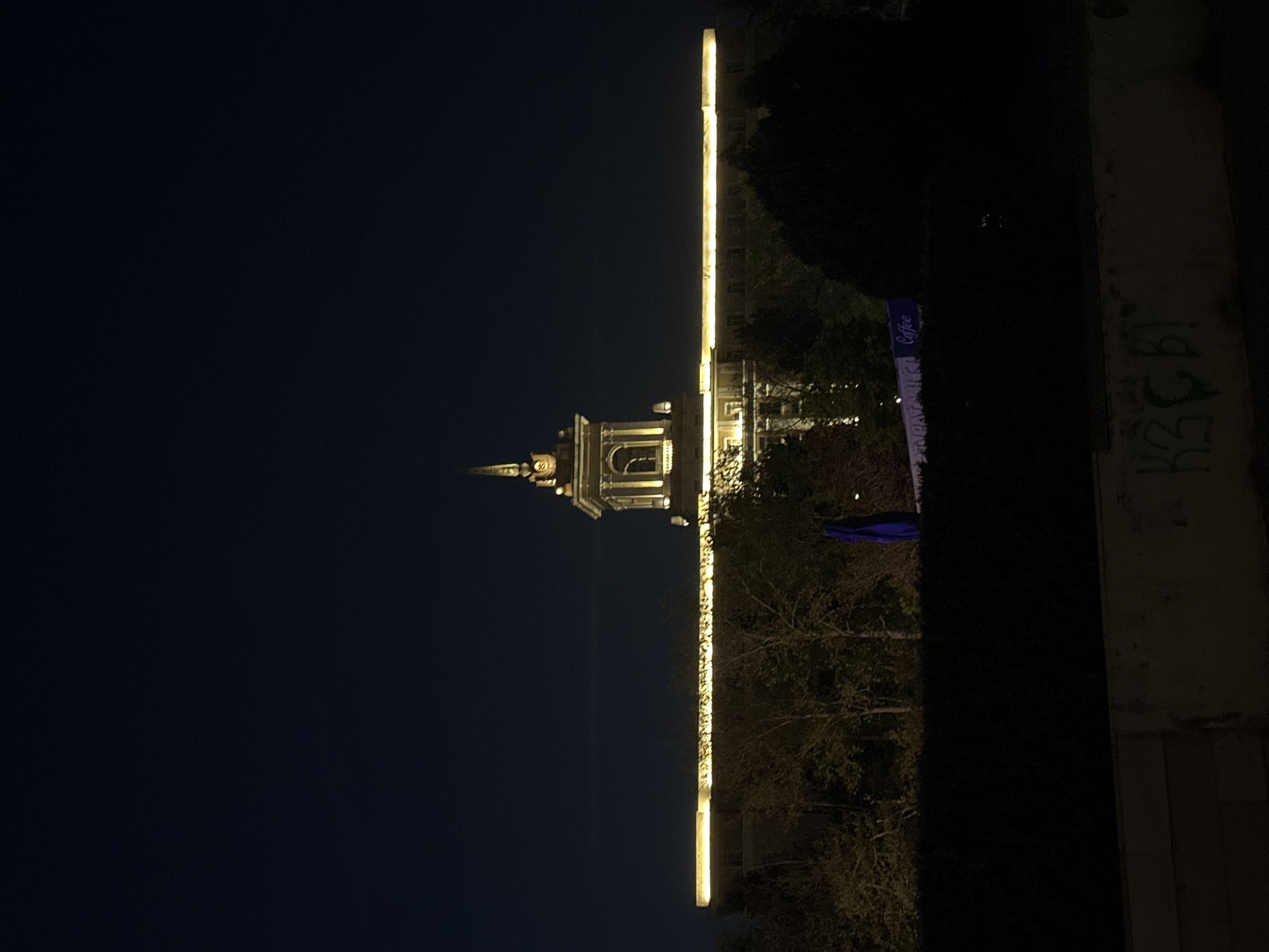 The image shows a building illuminated at night. Based on the architectural style, particularly the tower design, it is likely the Dormition of the Mother of God Cathedral in Varna, Bulgaria. The building is lit with warm, golden lights, contrasting with the dark night sky. To the right of the cathedral, some vegetation and a wall with graffiti can be seen. A banner with "Coffee" can also be seen.