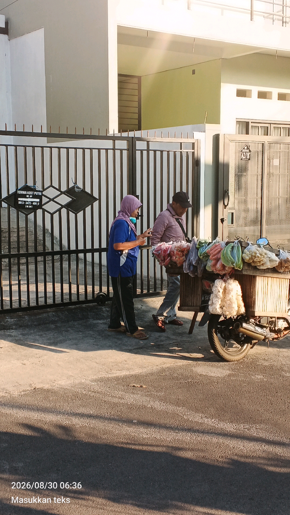 In Sumberporong, Indonesia, on a sunny day, a woman wearing a blue shirt, black pants, and a light purple hijab stands by a black metal gate. She is looking at her phone. Beside her, a man in a light-colored jacket and dark pants, wearing a black baseball cap, stands in front of a motorcycle-drawn cart filled with various packaged snacks and produce. The cart has a license plate that reads "N 507 Y". The background shows a building with a light green wall and white trim. The ground is paved, with shadows cast by the gate and the subjects. A date and time stamp at the bottom of the image indicates "2026/08/30 06:36" and the text "Masukkan teks".