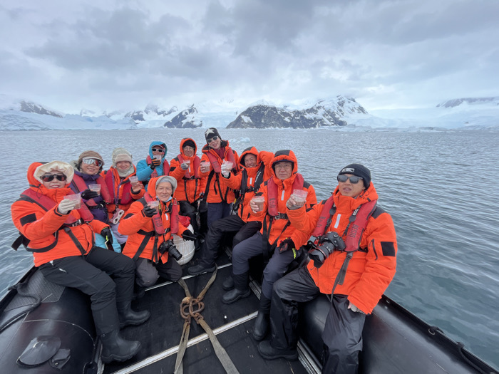 A group of ten people in bright orange life vests, seated in a dark inflatable boat on the water. Each person holds a small, clear cup. Snow-covered mountains and icebergs form a background under an overcast sky. The group is diverse in age and attire, with some wearing hats and sunglasses. The foreground is dominated by the boat and the water.