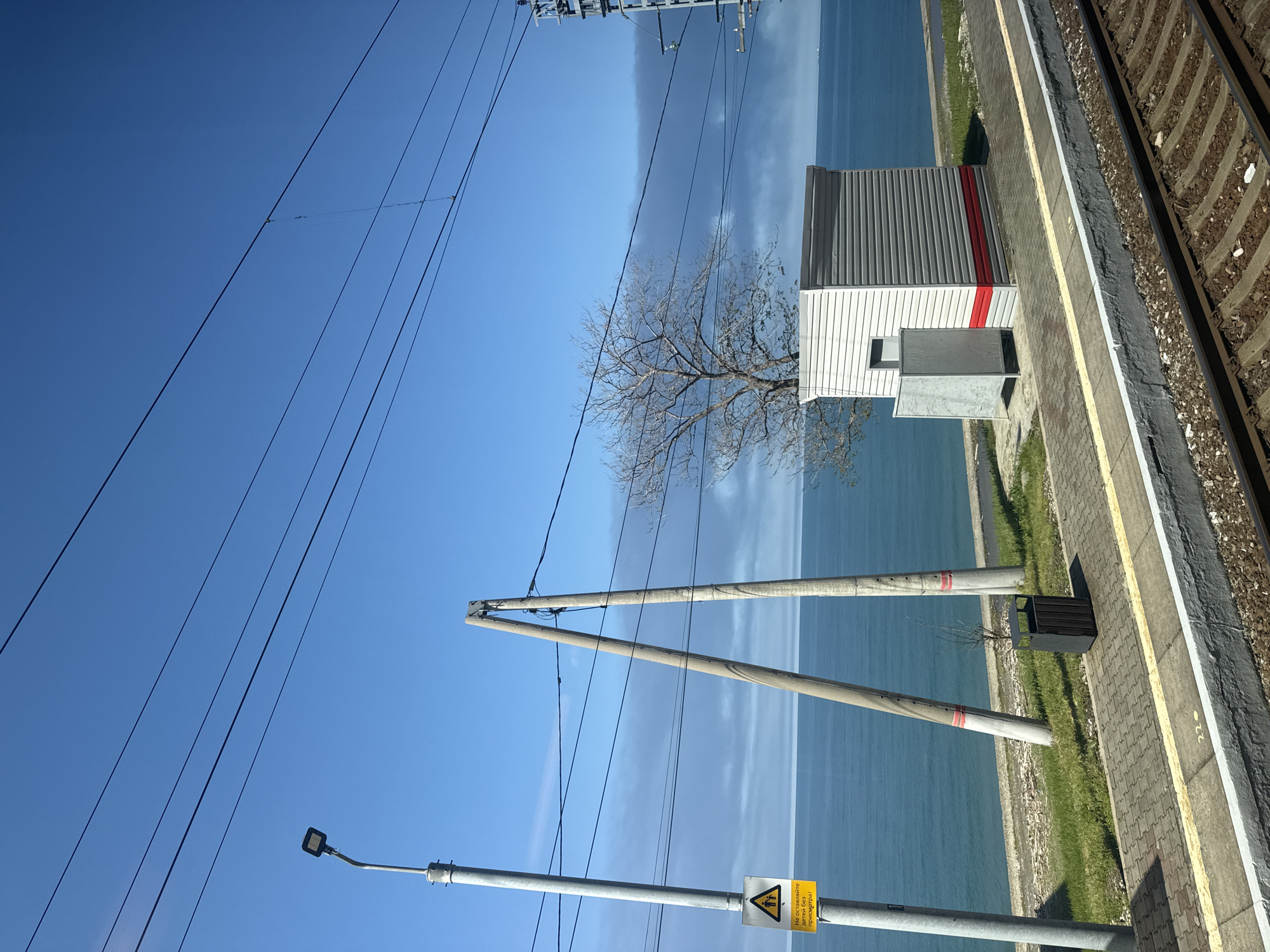 The image depicts a railway platform with train tracks on the right. Adjacent to the tracks is a yellow safety line and a paved platform with a green grassy area between the platform and the water. The water appears to be a lake or sea. On the platform, there is a small gray and white building with a red stripe. A bare tree stands near the building. Power lines run across the sky. There are two concrete poles with red stripes along the platform and a utility pole with a lamp and a yellow sign. The scene suggests a rural or semi-rural location, possibly near a body of water. The city and specific location are unknown.