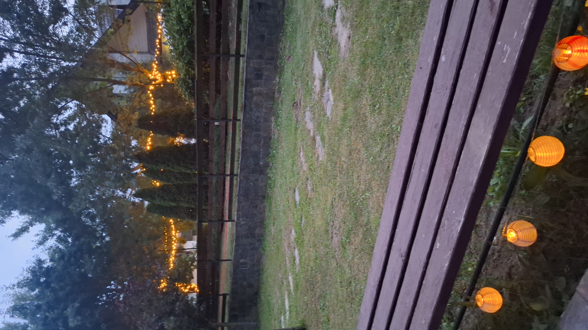 The image depicts an outdoor scene in Razlog, Bulgaria, likely in the evening or at dusk. The primary subjects are a wooden bench in the foreground, partially obscuring the view of a grassy area beyond. A line of decorative, illuminated orange paper lanterns extends along the edge of the bench, casting a warm glow.
In the background, trees with dense foliage are visible, some adorned with strings of yellow lights that create a festive ambiance. A wooden fence runs horizontally across the middle ground, separating the grassy area from a darker, possibly wooded or vegetated zone. Beyond the fence, a stone wall or structure is partially visible. The sky appears overcast or twilight, suggesting a dim natural light.
The scene evokes a sense of a relaxed outdoor gathering or a tranquil evening setting. There are no people visible in the image. The overall atmosphere is peaceful, enhanced by the warm lighting from the lanterns and the string lights. No text is visible.