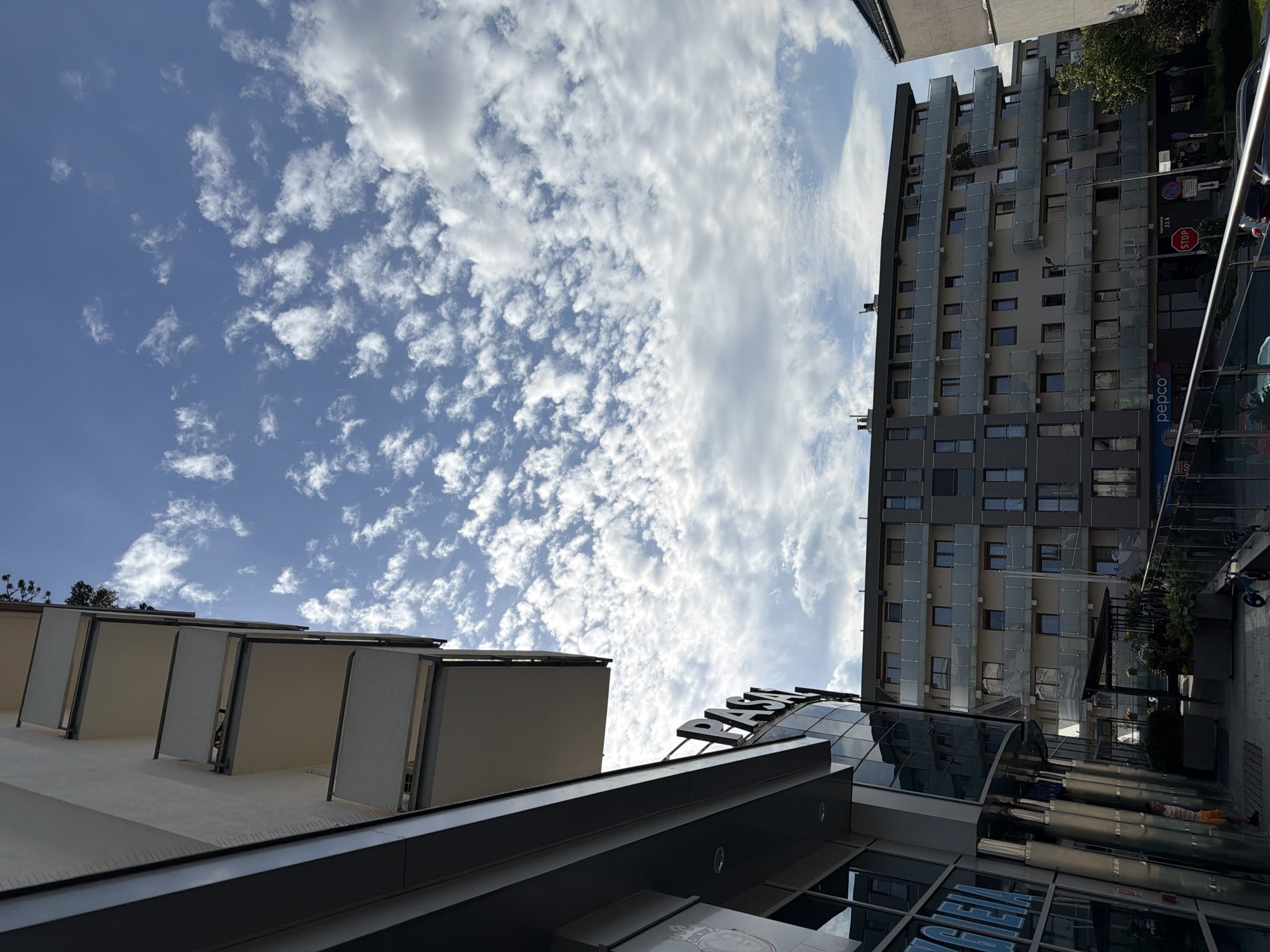 An eye-level, exterior shot captures a low-angle perspective of modern architecture against a partly cloudy blue sky. In the foreground to the left, a portion of a building with a curved, reflective glass facade is visible, displaying blue text that reads "CIEJA". Above this, a white building with a series of balconies extends upwards. To the right of the frame, a multi-story residential building with numerous balconies and windows dominates the mid-ground. A street with parked cars, a "stop" sign, and various other traffic signs is visible at the base of this building. The sky is filled with scattered, white, cumulus clouds.