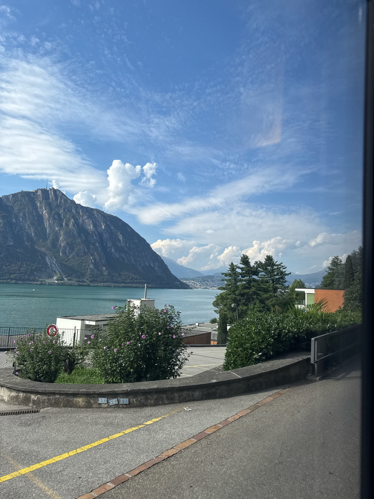 The image captures a scenic view of Lake Lugano with a large mountain dominating the left side of the frame. In the foreground, a paved road curves to the right, marked with a yellow line and a brick border. A low concrete wall, topped with a stone coping, lines the curve of the road and encloses a small grassy area with some bushes and purple flowers. Two metallic plaques, one reading "47" and the other "49 51", are affixed to the wall. Behind the wall, a white building with a flat roof and an external chimney stands near the lake. Several evergreen trees and shrubs are visible in the middle ground. Further back, more buildings and a brick-colored structure are nestled amongst dense greenery. The lake itself is a vibrant turquoise, and on its surface, a small white boat is visible. The background features rolling hills and a distant town. The sky above is a clear blue, punctuated by scattered white clouds, some of which are large and puffy, while others are thin and wispy. The image appears to be taken from an elevated position, possibly a road or pathway leading downwards towards the lake. The overall impression is one of a tranquil, sunlit day in a picturesque lakeside town, likely Bissone, Switzerland, given the geological features and architectural style.