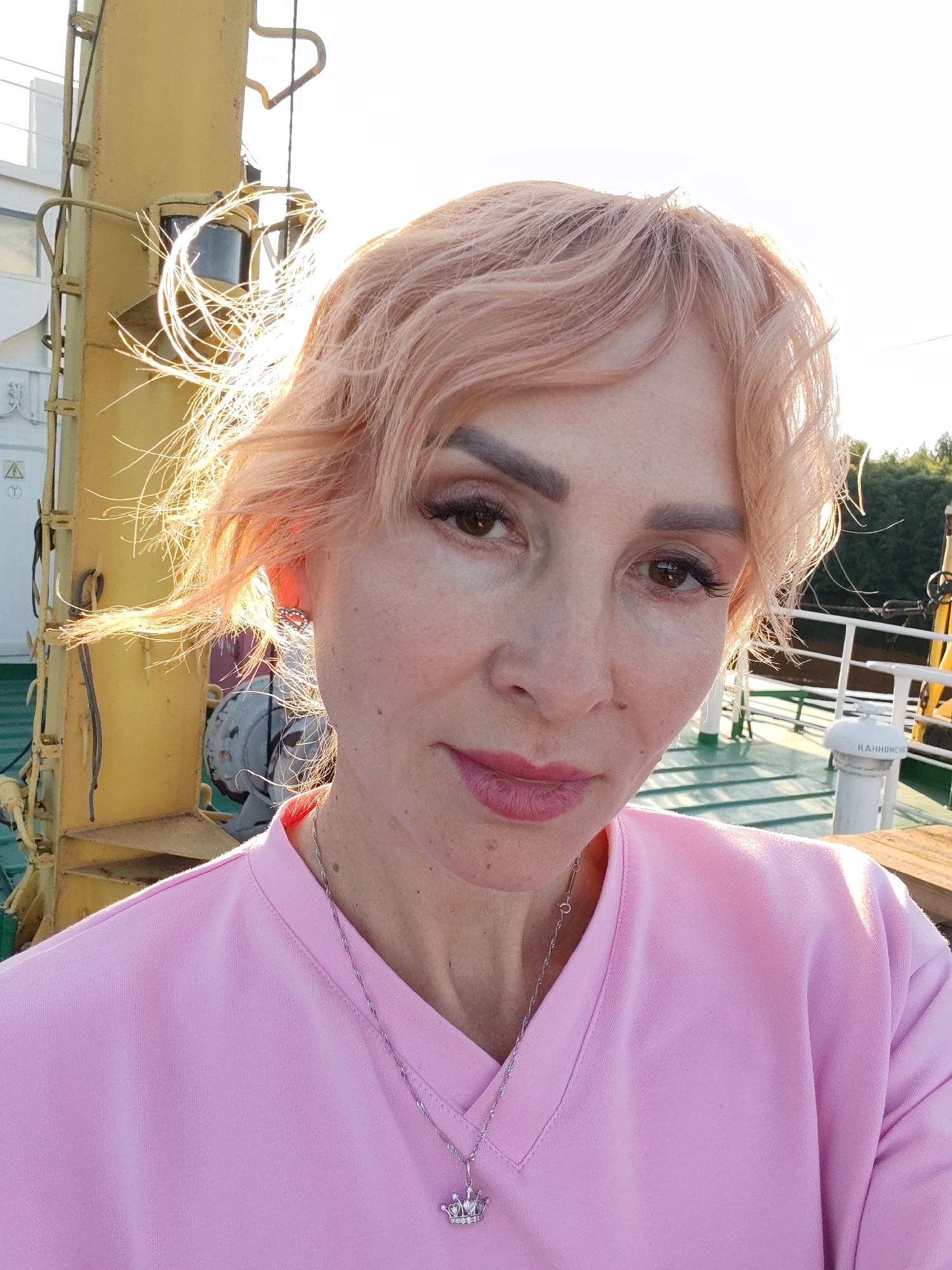 A medium shot captures a Caucasian woman with short, peachy-pink hair and light skin. She is wearing a pink V-neck top and a silver necklace with a crown pendant. Her eyes are brown, and she has visible makeup, including eyeliner and mascara. The background suggests an outdoor setting, possibly on a boat or a dock, with a yellow structure to the left, greenery in the distance, and a railing with white posts. The lighting indicates it is daytime, with the sun creating a flare around her hair. The location is identified as Kurdiug, Russia.