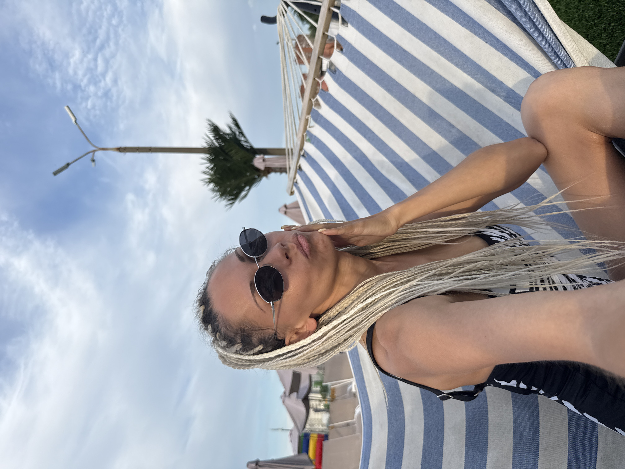 In Rusanovo, Russia, a woman with blonde braids and sunglasses rests in a hammock. The hammock has blue and white stripes and is outdoors, with a palm tree and a lamppost visible in the background under a cloudy sky. She is wearing a black and white swimsuit.