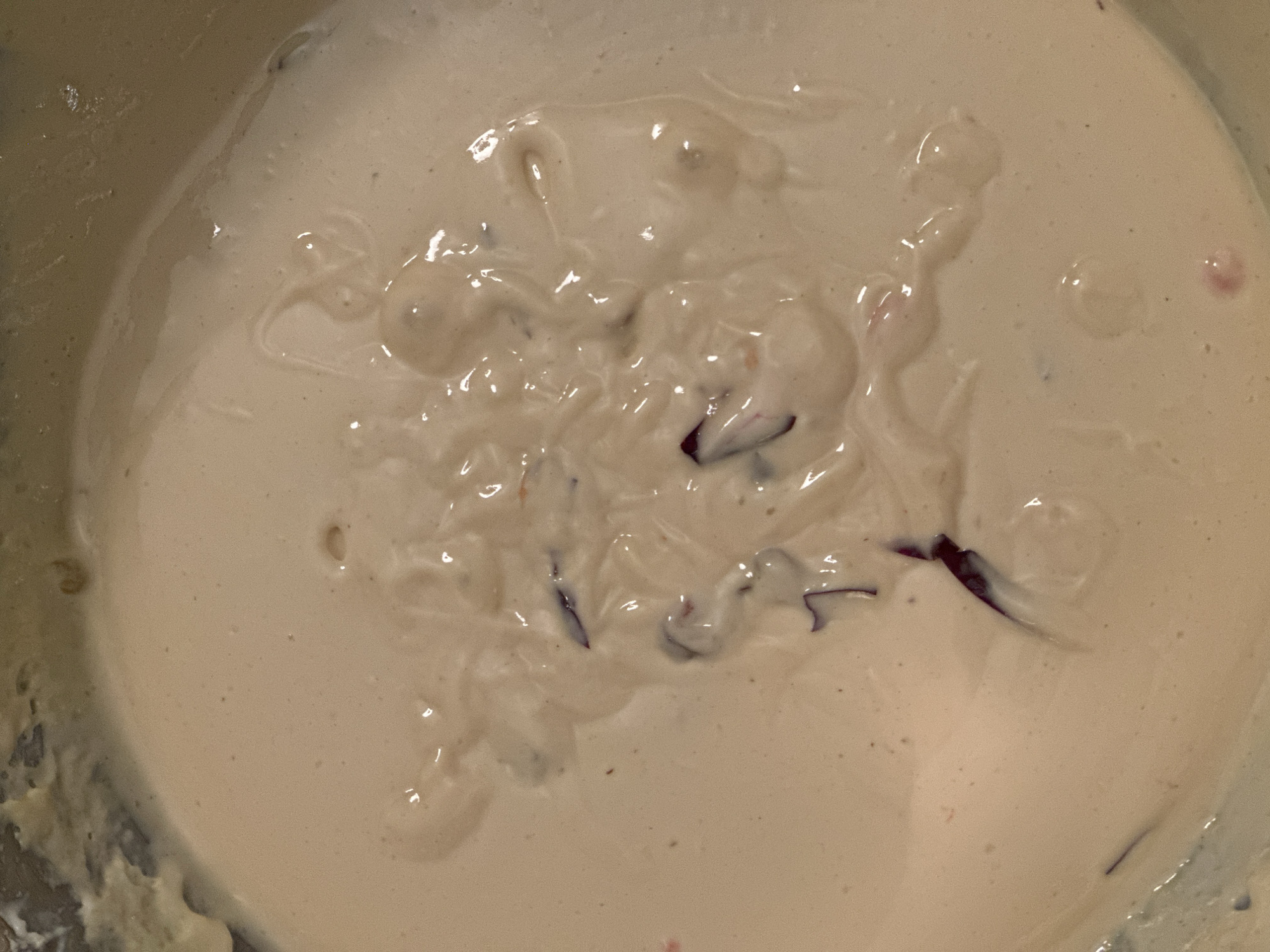The image is a close-up, overhead shot of a creamy, pale beige substance with darker purple and pinkish-red flecks mixed throughout. The texture is smooth and viscous, with some areas showing ripples and glossy highlights, suggesting it is a liquid or semi-liquid food item, possibly batter or a dessert. Some dark, elongated fragments, appearing stringy or petal-like, are visible within the mixture. There is no discernible information within the image to indicate a specific location in Nevinnomyssk, Russia.