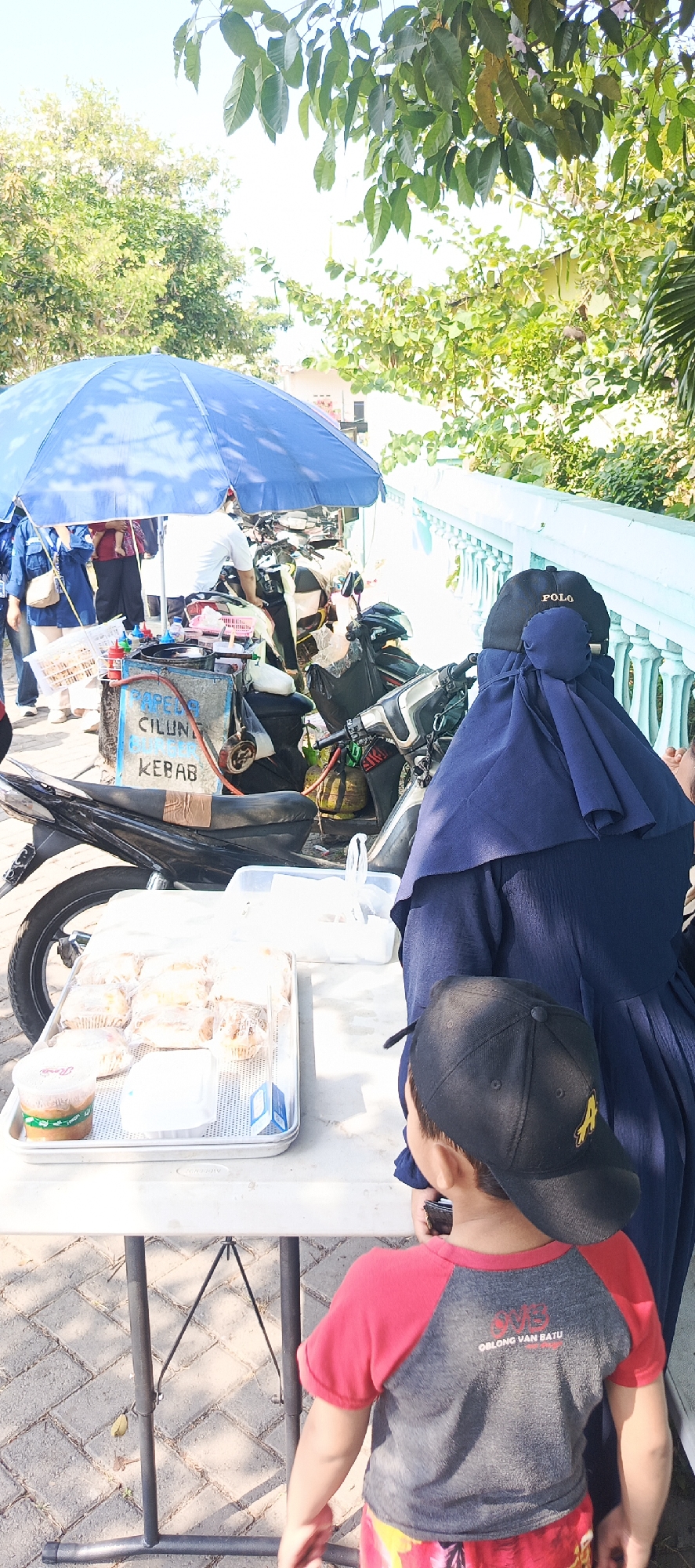 In Wono Plintahan, Indonesia, an outdoor scene depicts a street food stall under a large blue umbrella. The stall, decorated with a sign advertising "CILUN" and "KEBAB," features motorcycles and a gas tank as part of its setup. A tray of pastries is displayed on a white folding table in the foreground. Several individuals are present: a child wearing a black baseball cap and a red and grey t-shirt stands facing away from the camera, looking towards the stall. A woman in a dark blue hijab and dress is also visible. Other people are partially seen behind the stall and umbrella. The ground is paved with cobblestones. Lush green trees and foliage frame the top and right sides of the image, with a light blue railing visible in the background. The lighting suggests it is a sunny day.